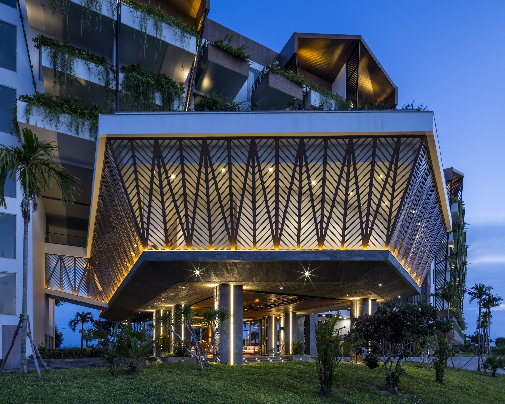 Cantilevered porte-cochère with backlit chevron-patterned metal screens and planted terraces above at blue hour