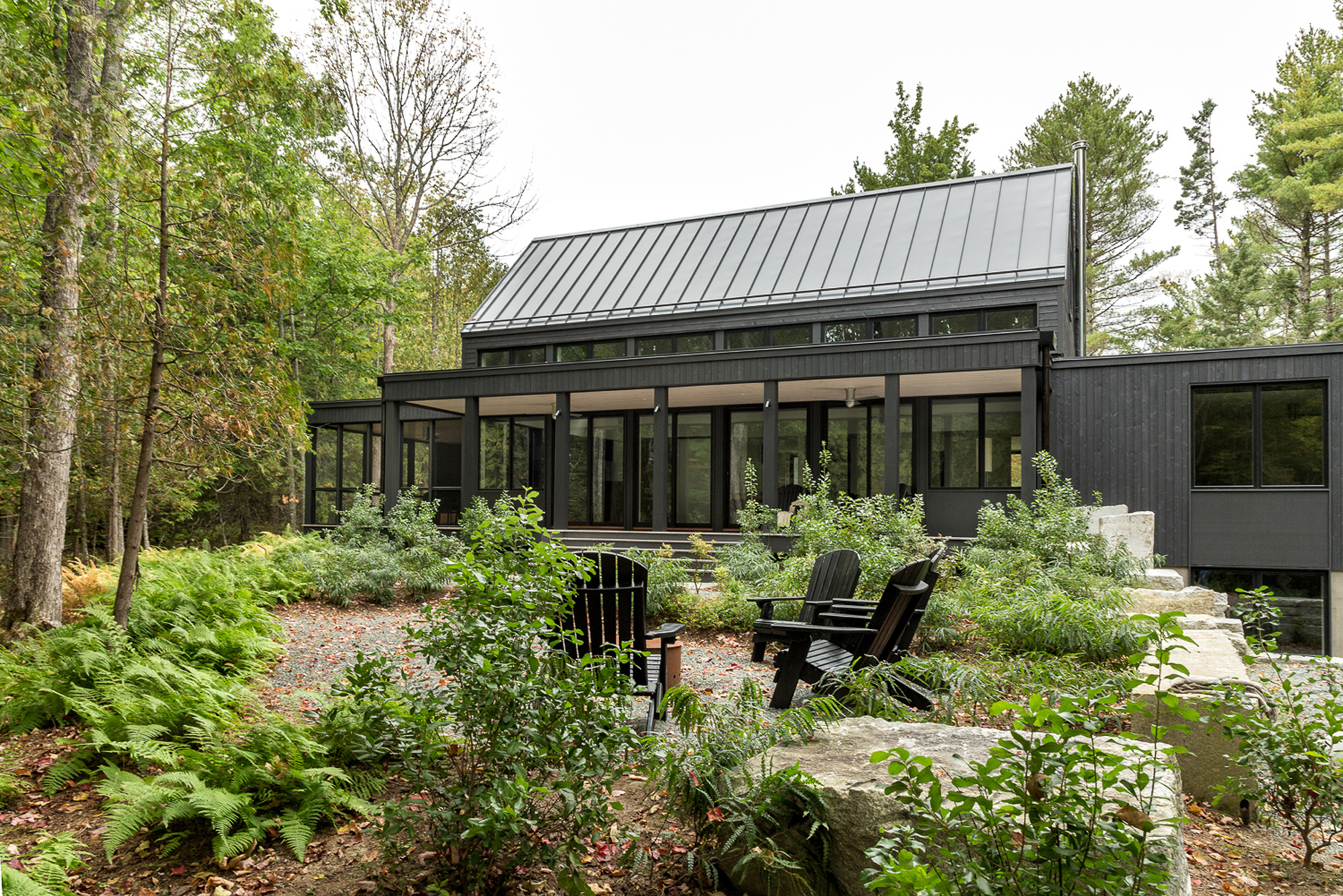Rear elevation with floor-to-ceiling windows opening to a patio with Adirondack chairs and naturalized plantings
