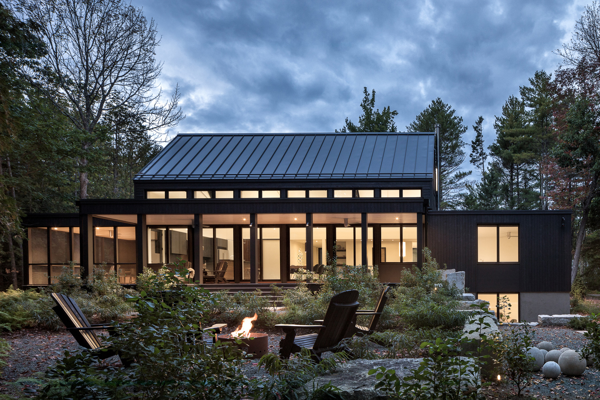 Rear elevation showing standing seam metal roof, glazed openings, and fire pit at twilight