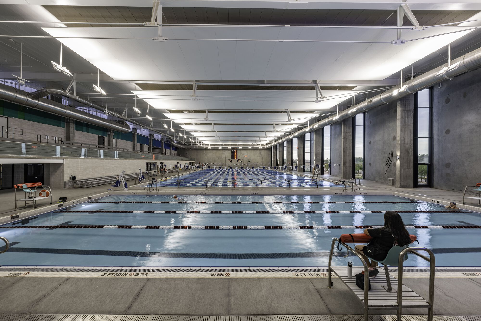 Olympic-length indoor swimming pool with concrete walls, exposed ceiling trusses and lane dividers
