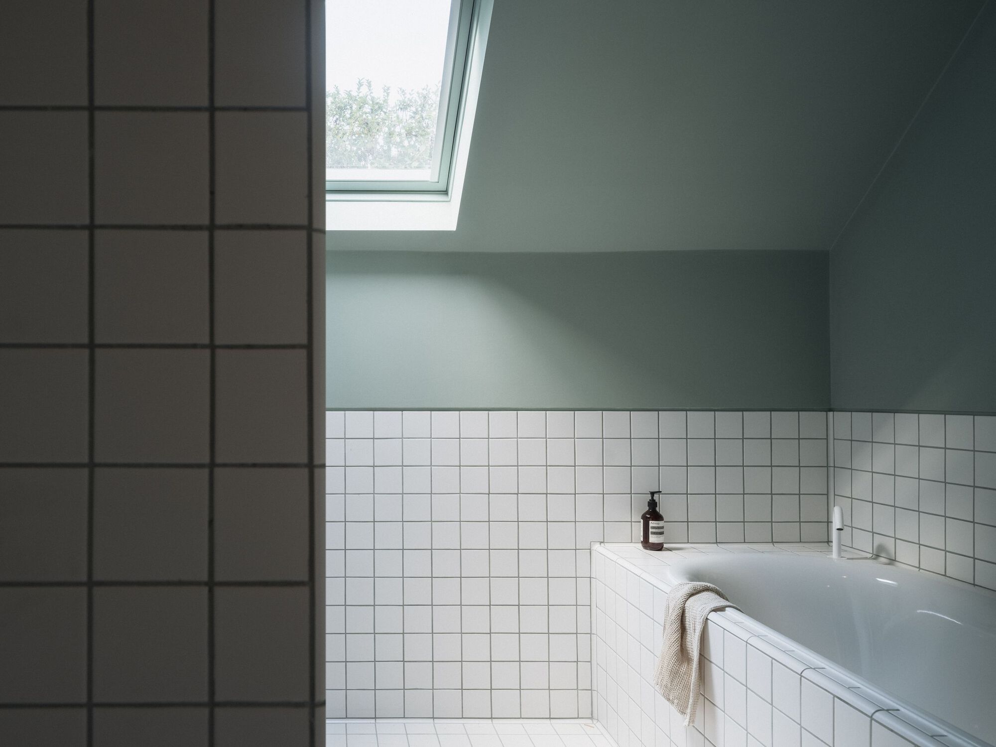 Bathtub corner with white square tiles under a sloped ceiling and angled skylight casting soft daylight