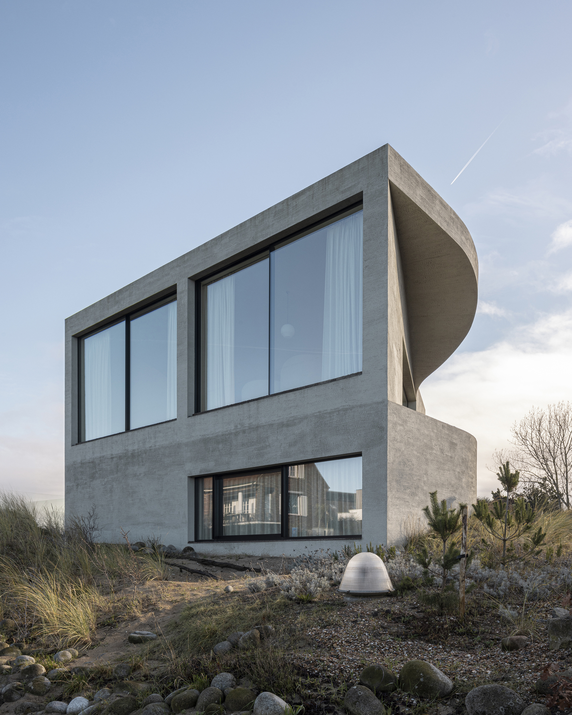 Rear facade at dusk with large glazed openings and cylindrical roof element above planted dunes