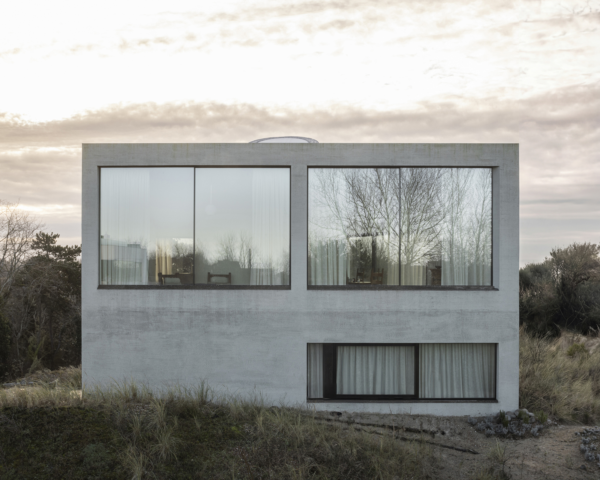 Rear facade with paired horizontal windows overlooking coastal dunes under overcast sky