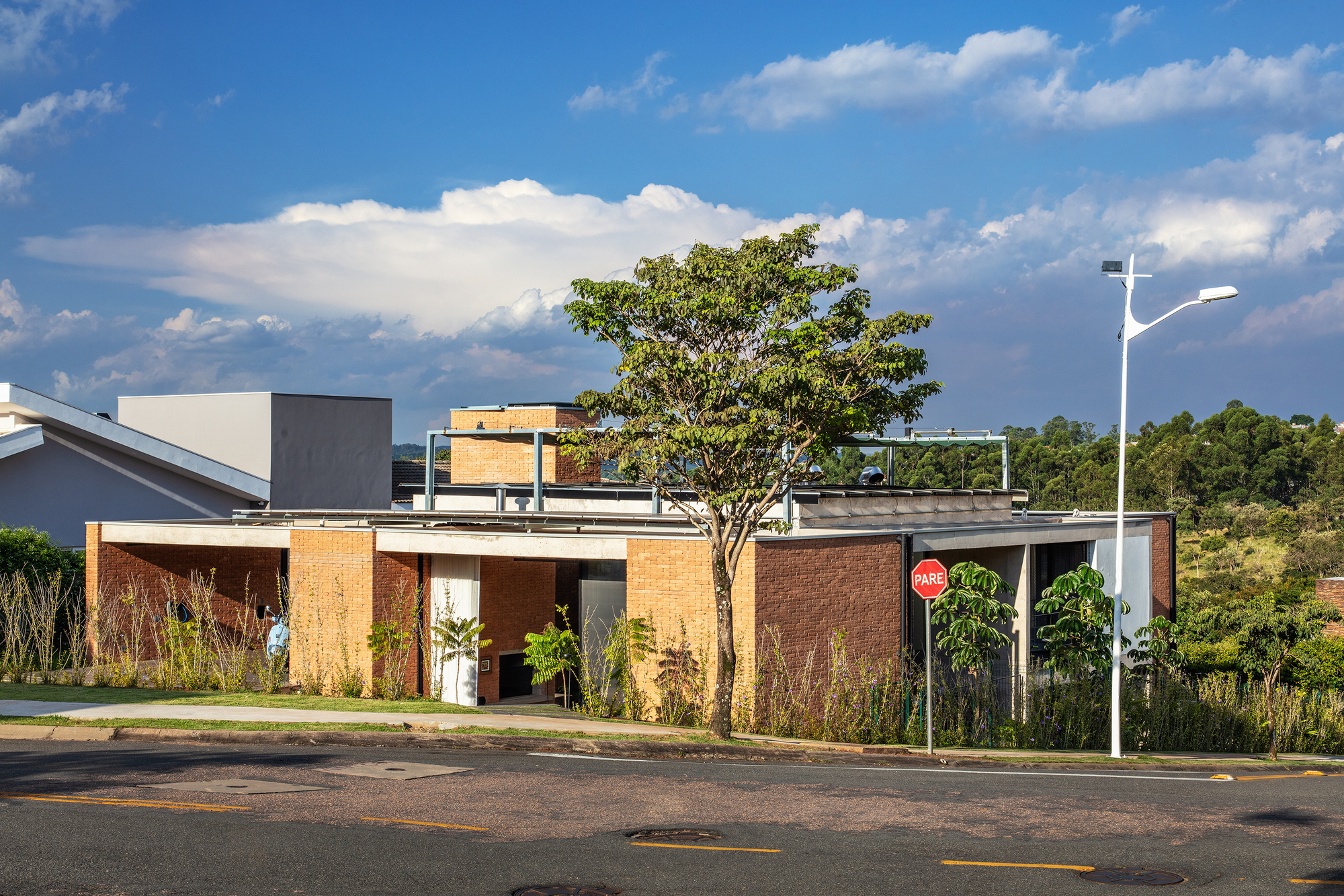 Street view of the low-slung residence with brick walls, planted garden beds and rooftop terrace under scattered clouds