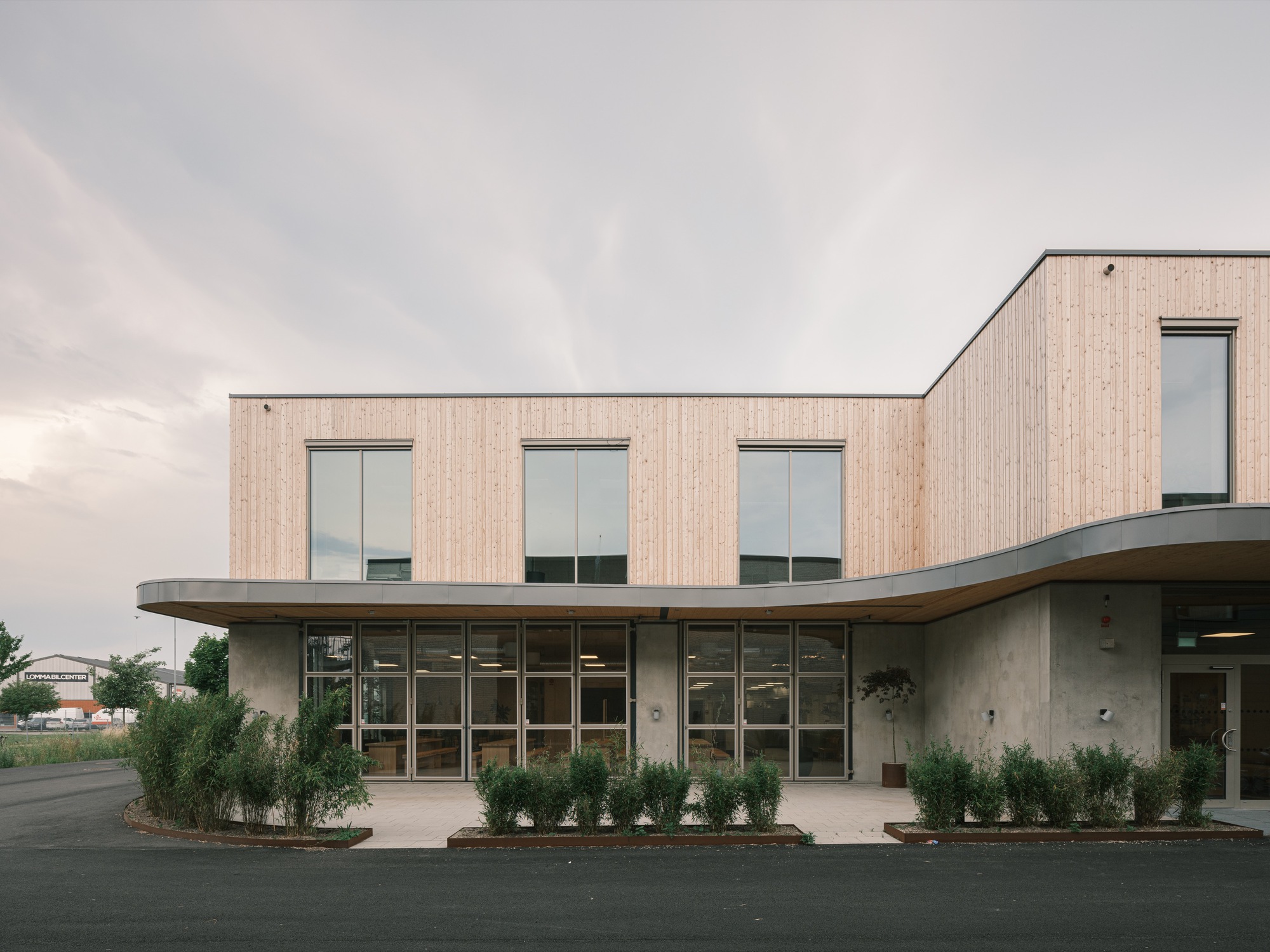 Two-storey facade with vertical timber cladding above a curved concrete base with planted beds at dusk