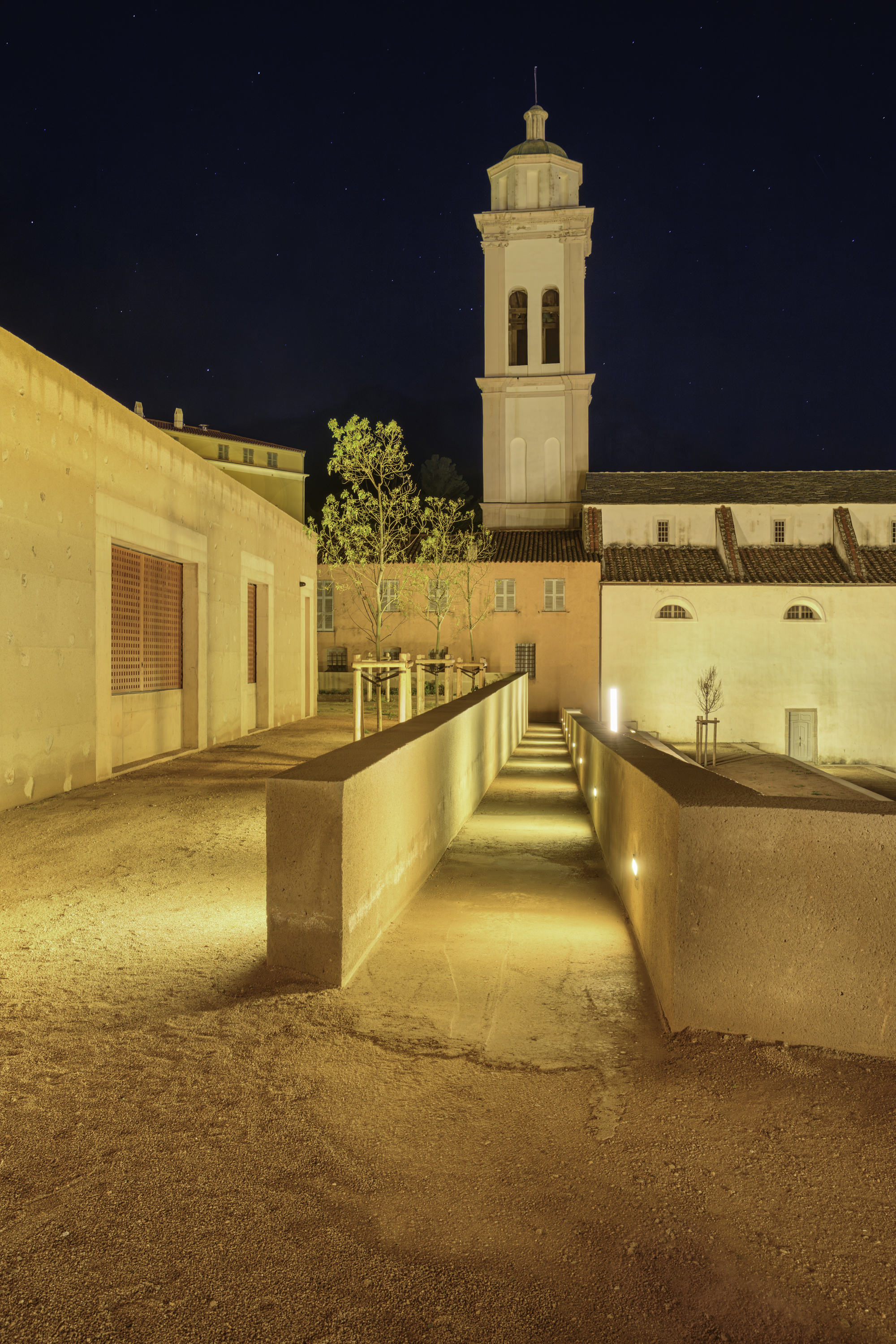 Night view of the illuminated courtyard passage with concrete walls framing the bell tower under a dark sky
