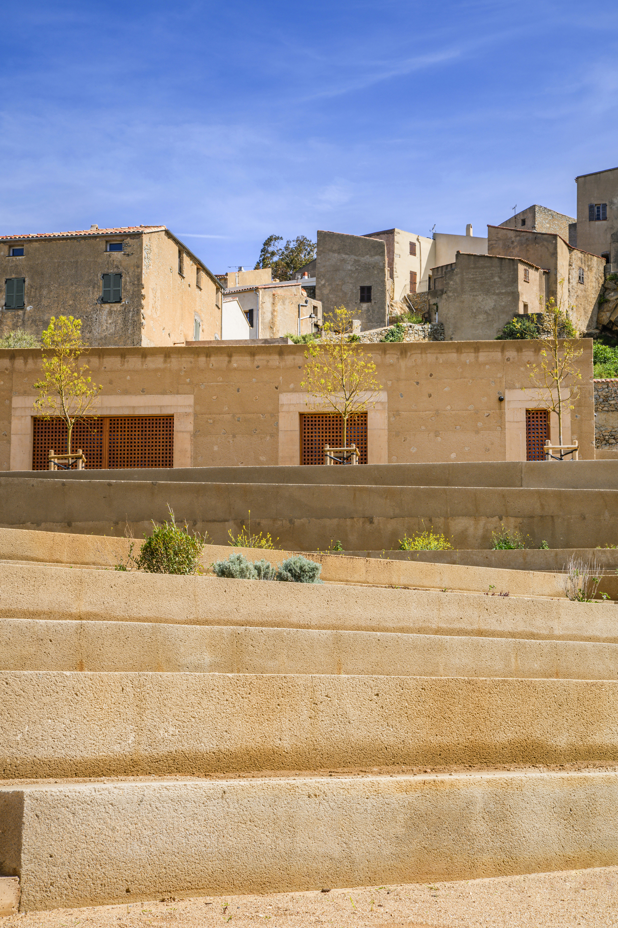 Stepped plaza with young trees and wood-slatted openings in the concrete retaining wall below the old village