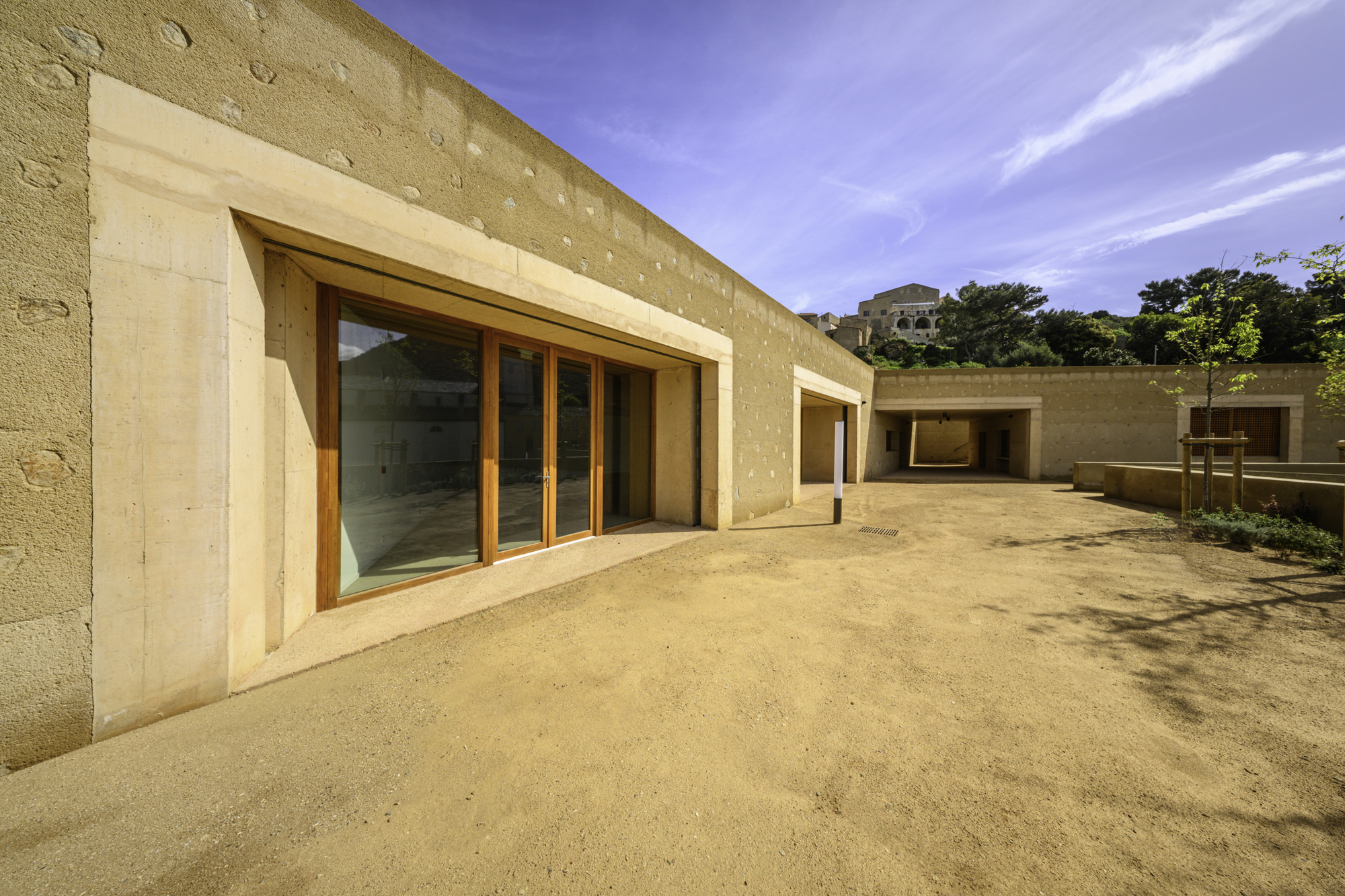 Exterior courtyard with rammed earth walls and timber-framed glazing under a blue afternoon sky