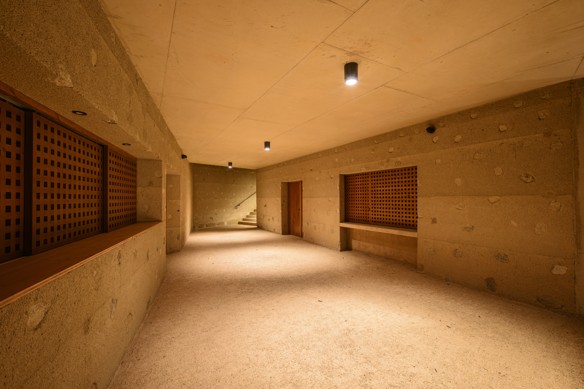 Basement corridor with rammed earth walls and timber lattice screens leading to a staircase