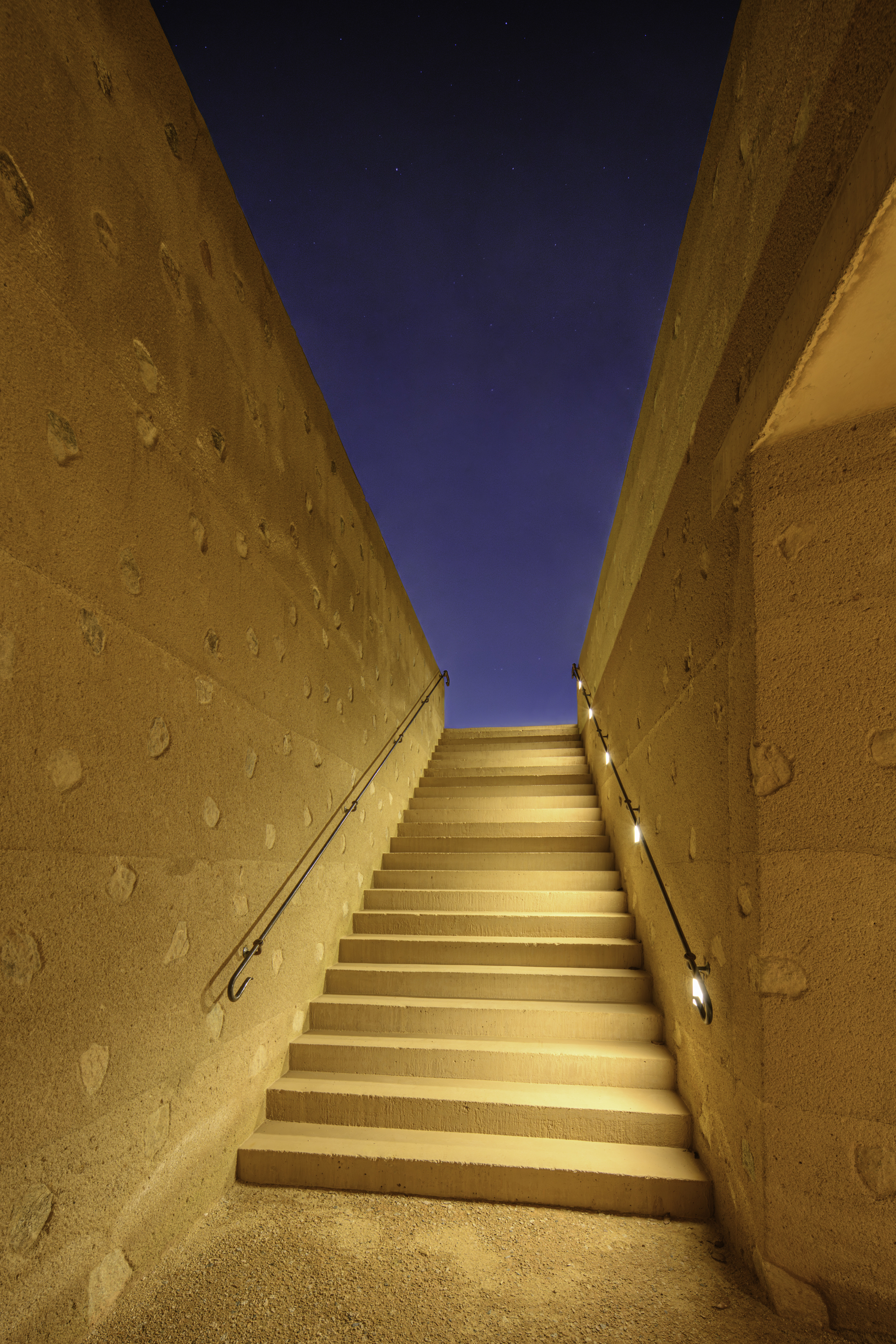 Concrete staircase ascending between rammed earth walls toward a narrow opening framing the night sky