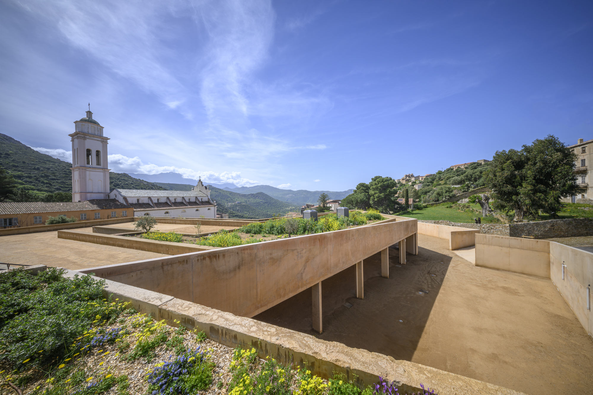 Elevated concrete roof terrace with planted beds alongside a historic church tower and mountains