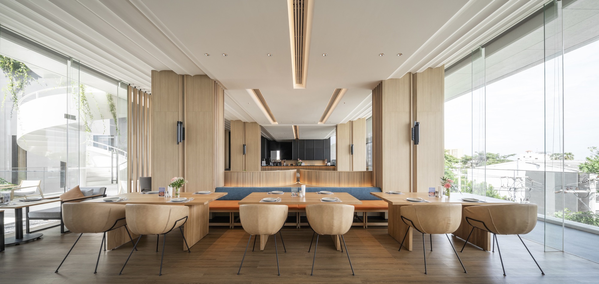 Communal dining space with long timber table, upholstered seating, and vertical wood paneling framing the kitchen beyond
