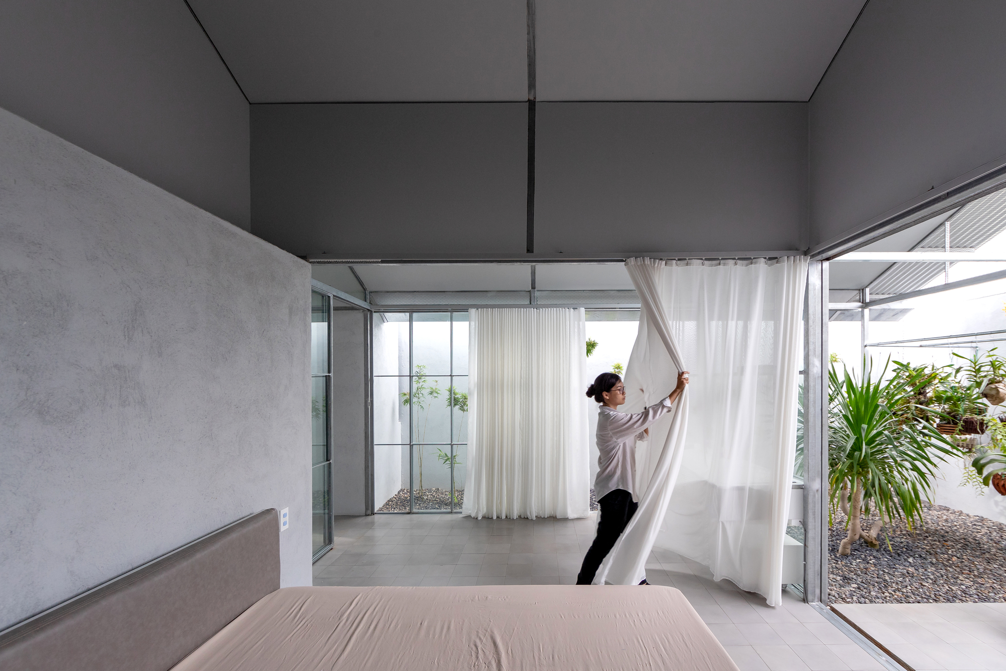 Double-height bedroom with concrete walls and person adjusting white curtain beside glazed courtyard