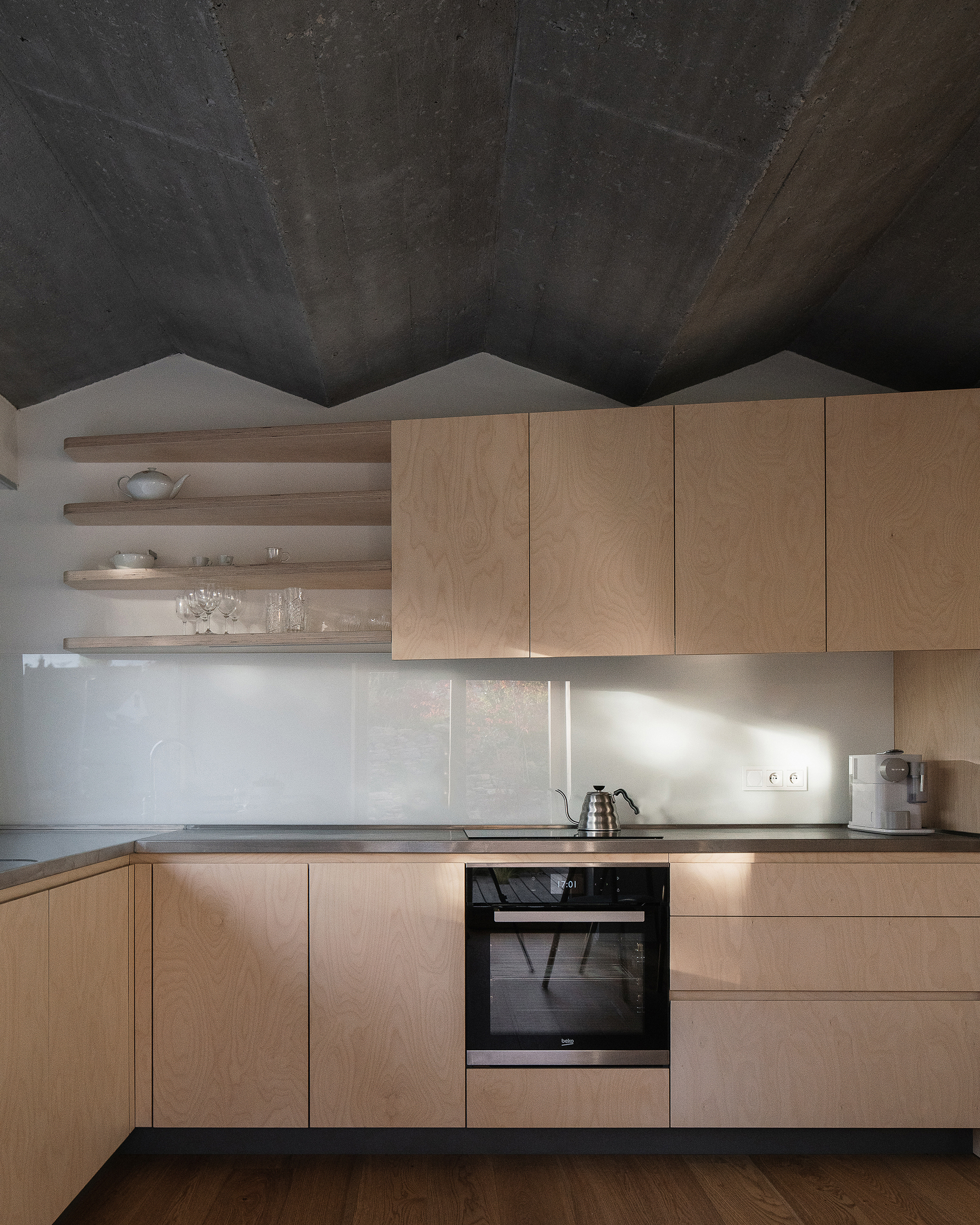 Light wood kitchen cabinetry with open shelving under a folded concrete ceiling catching afternoon sunlight
