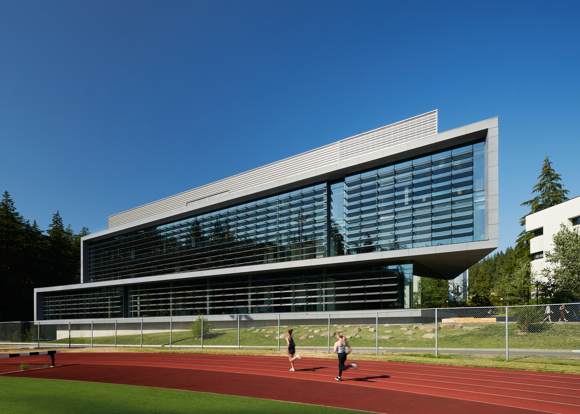 Horizontal louver facade and cantilevered upper volume viewed from the adjacent running track under a clear sky