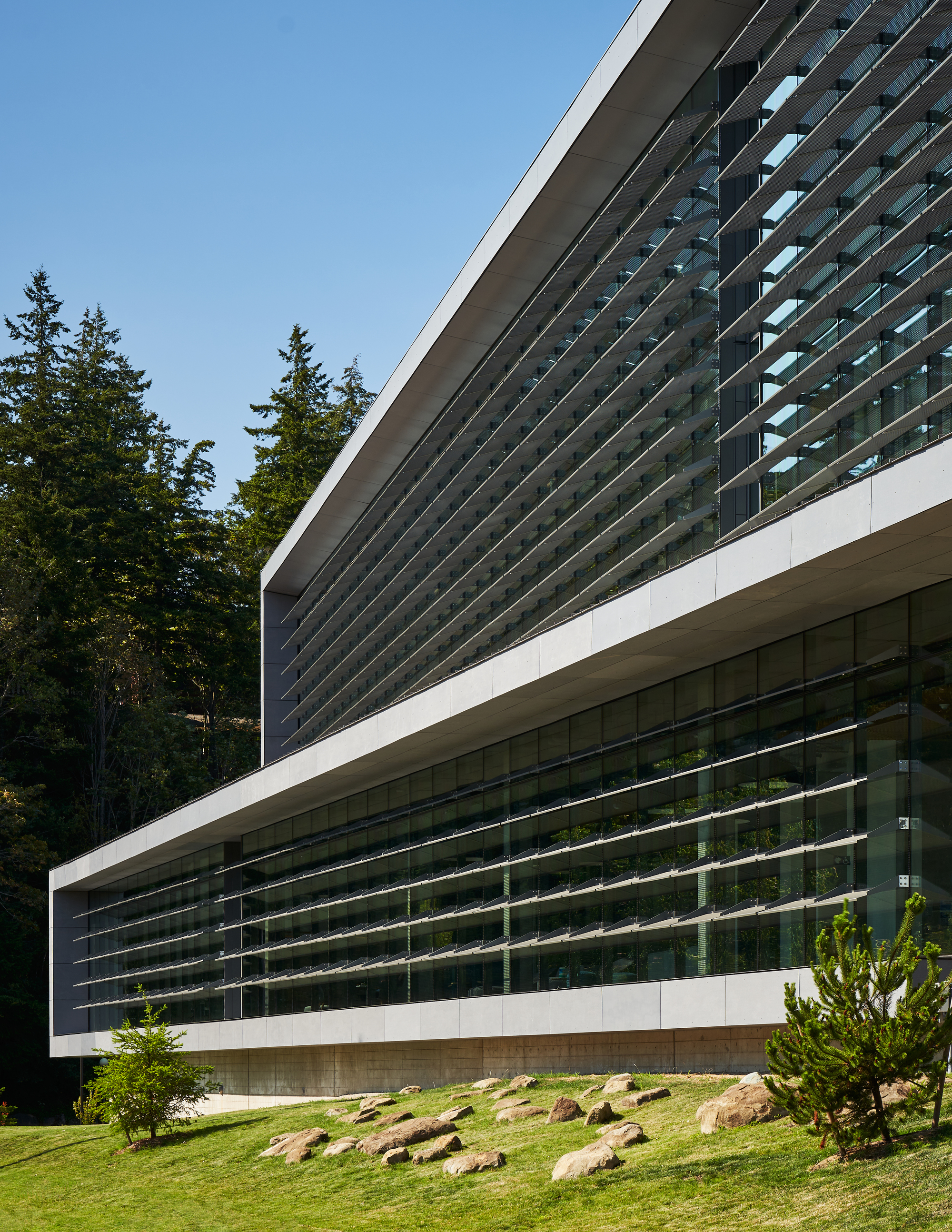 Layered horizontal louver system wrapping the building facade above a landscaped slope with scattered boulders