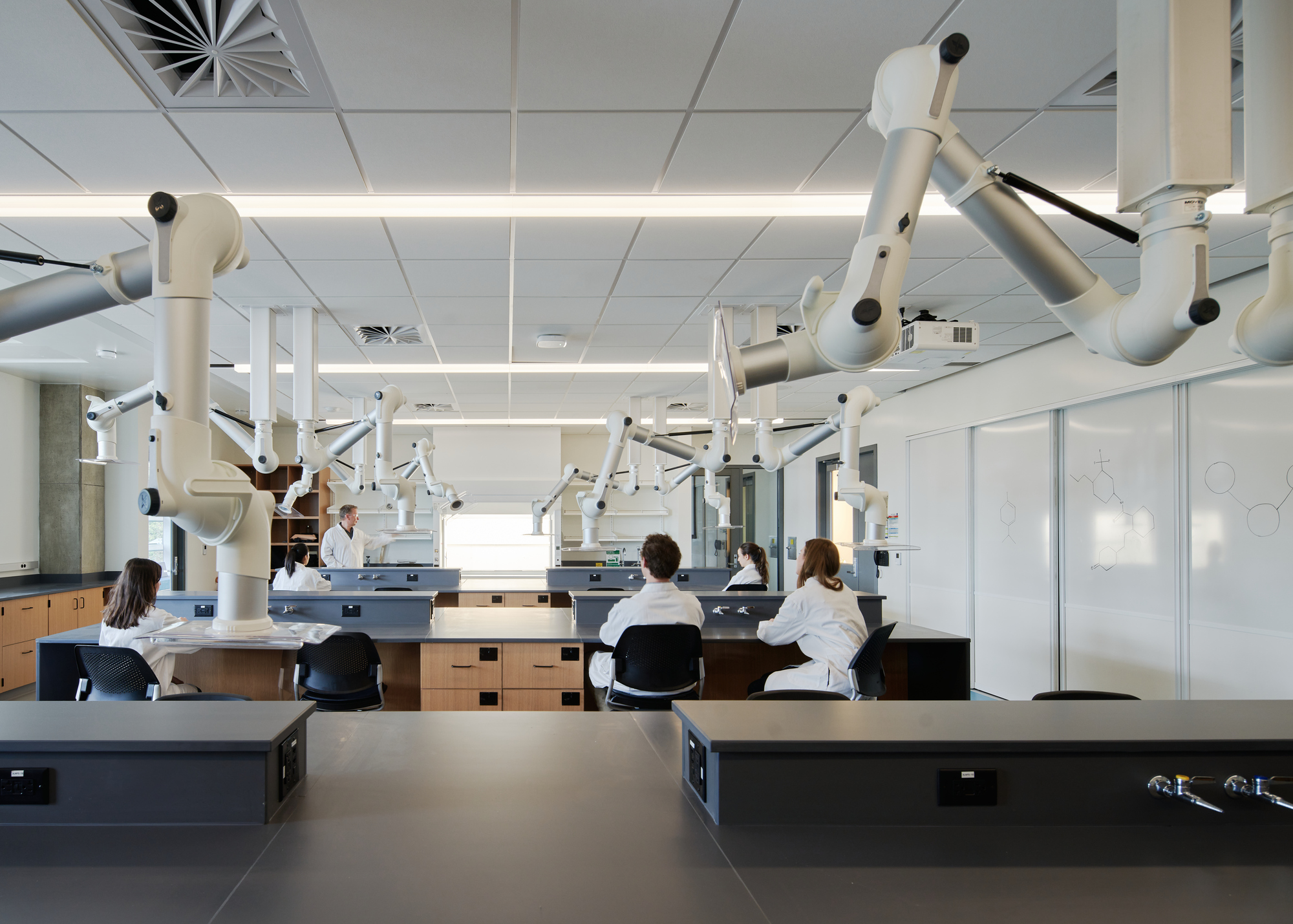 Laboratory workspace with rows of articulated microscope arms and students in white coats at workbenches