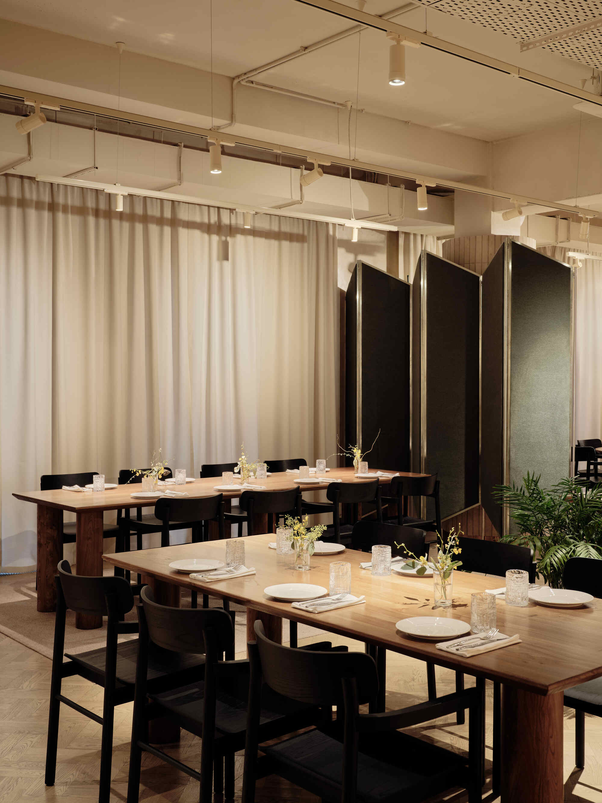 Two timber dining tables with black chairs separated by black folding screens and sheer curtains