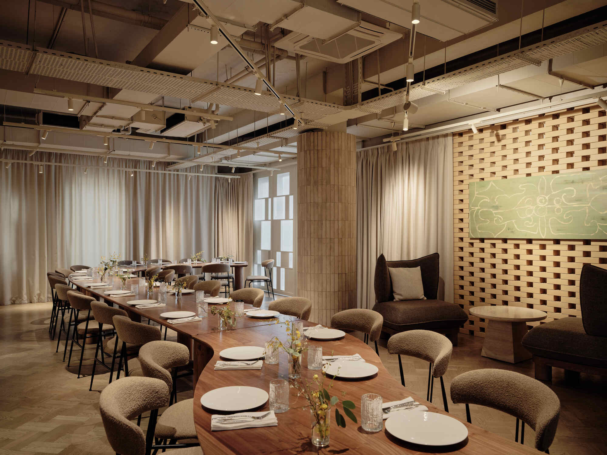 Long dining tables set with white plates beneath exposed ceiling ducts and sheer curtains filtering daylight