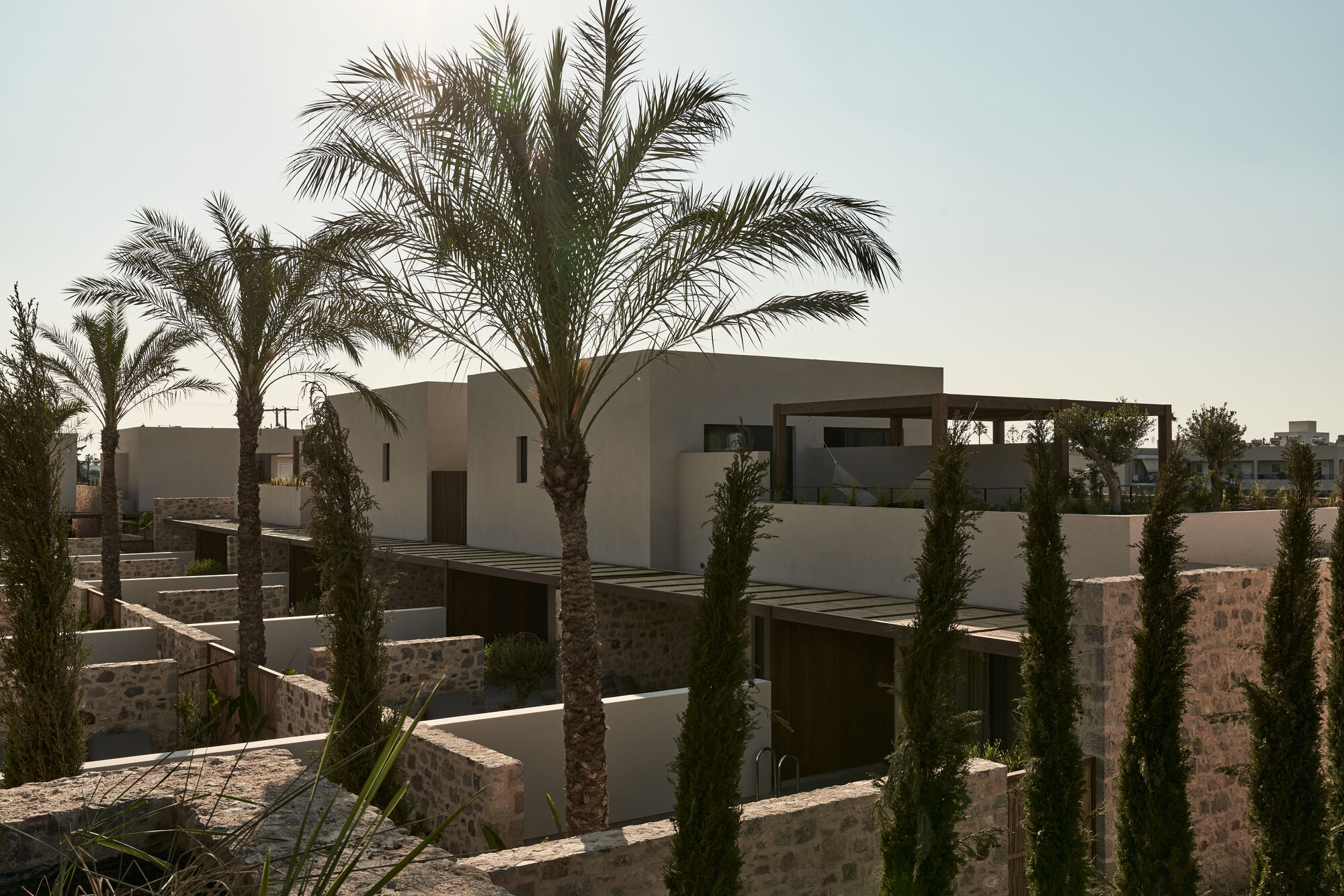 Clustered residential volumes with stone base walls and smooth stucco upper floors among cypresses and palms