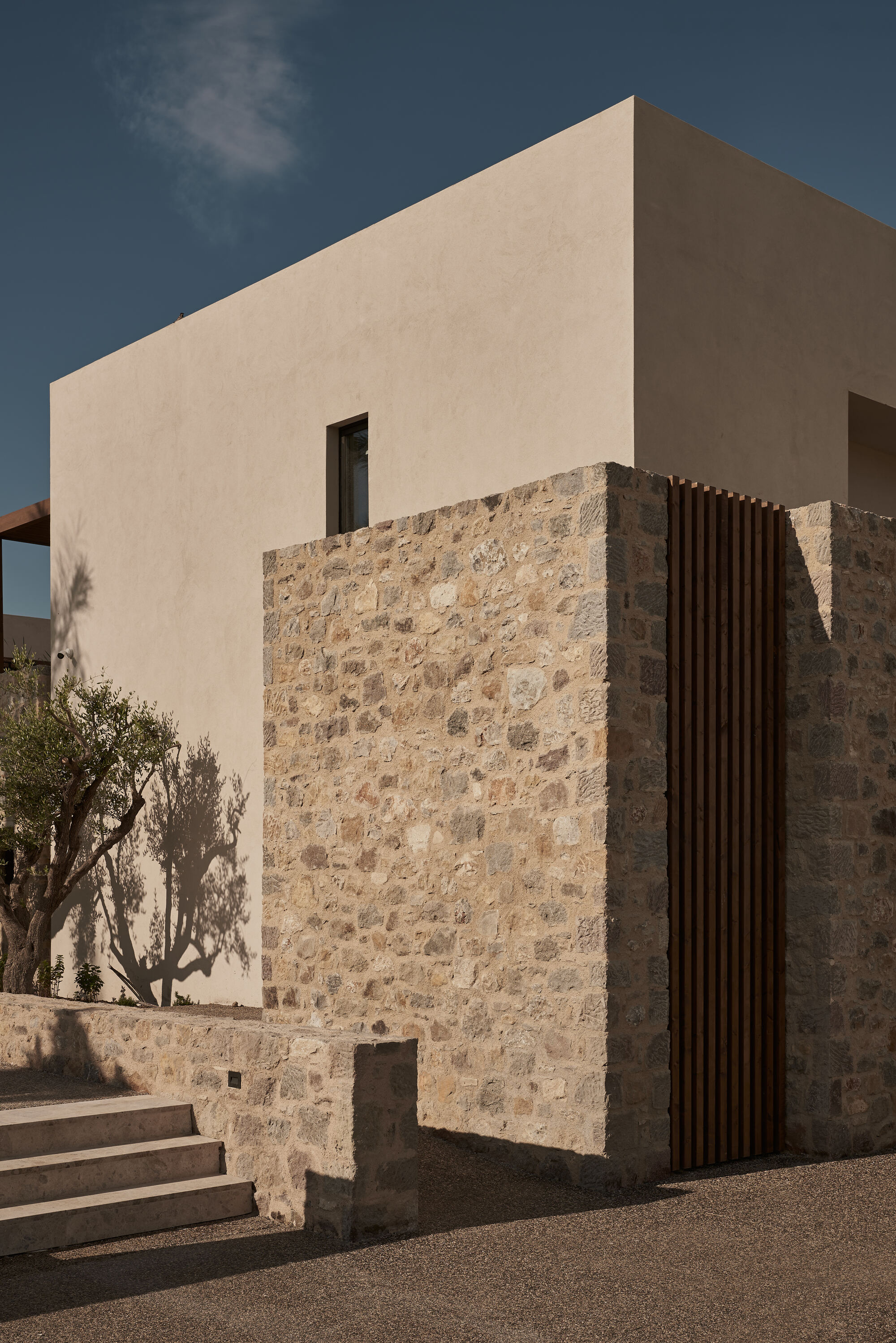 Corner facade combining pale stucco upper volume with rough stone lower wall and vertical timber screen