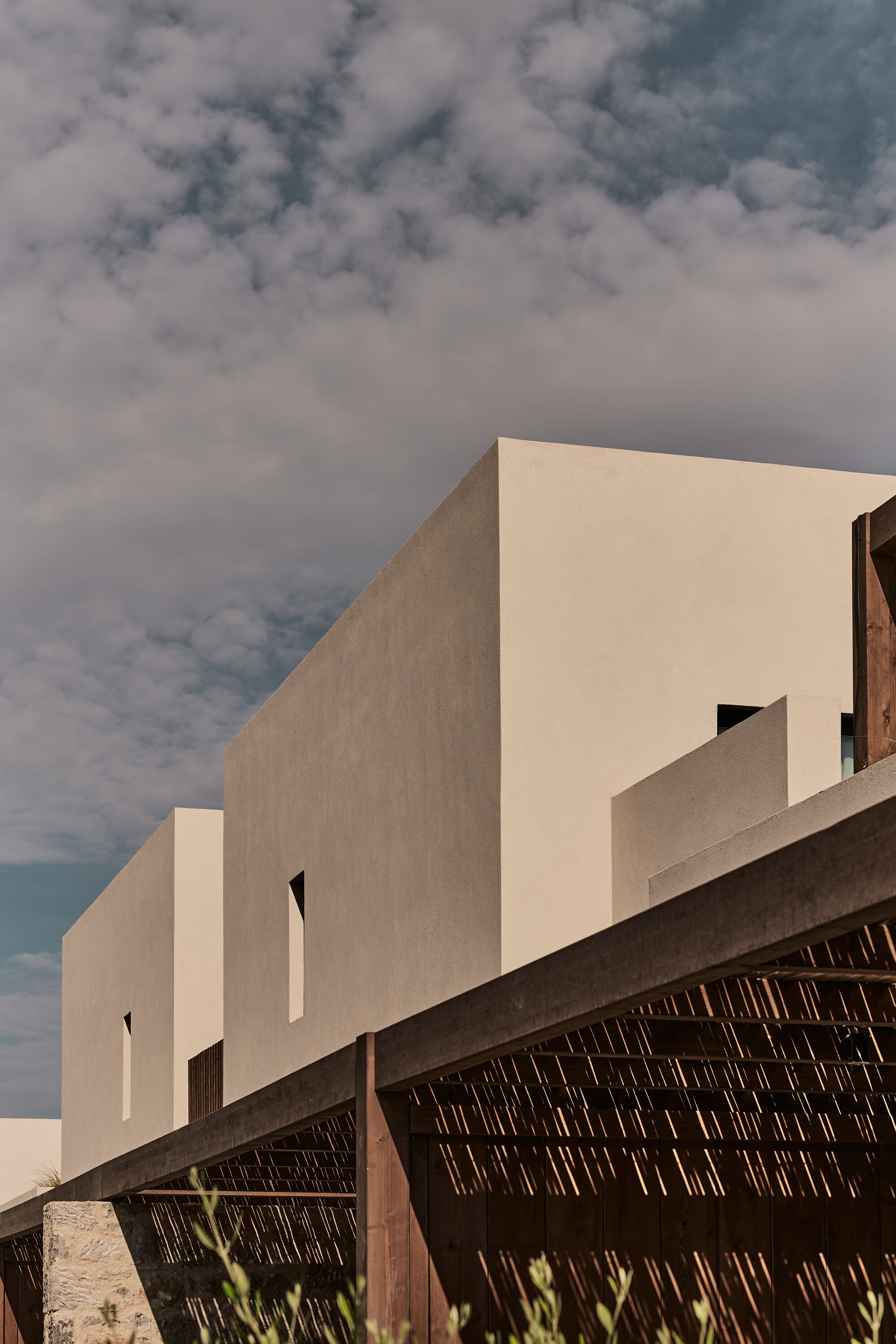 Stucco volumes with tall slot windows rising above a woven timber screen under cloudy skies