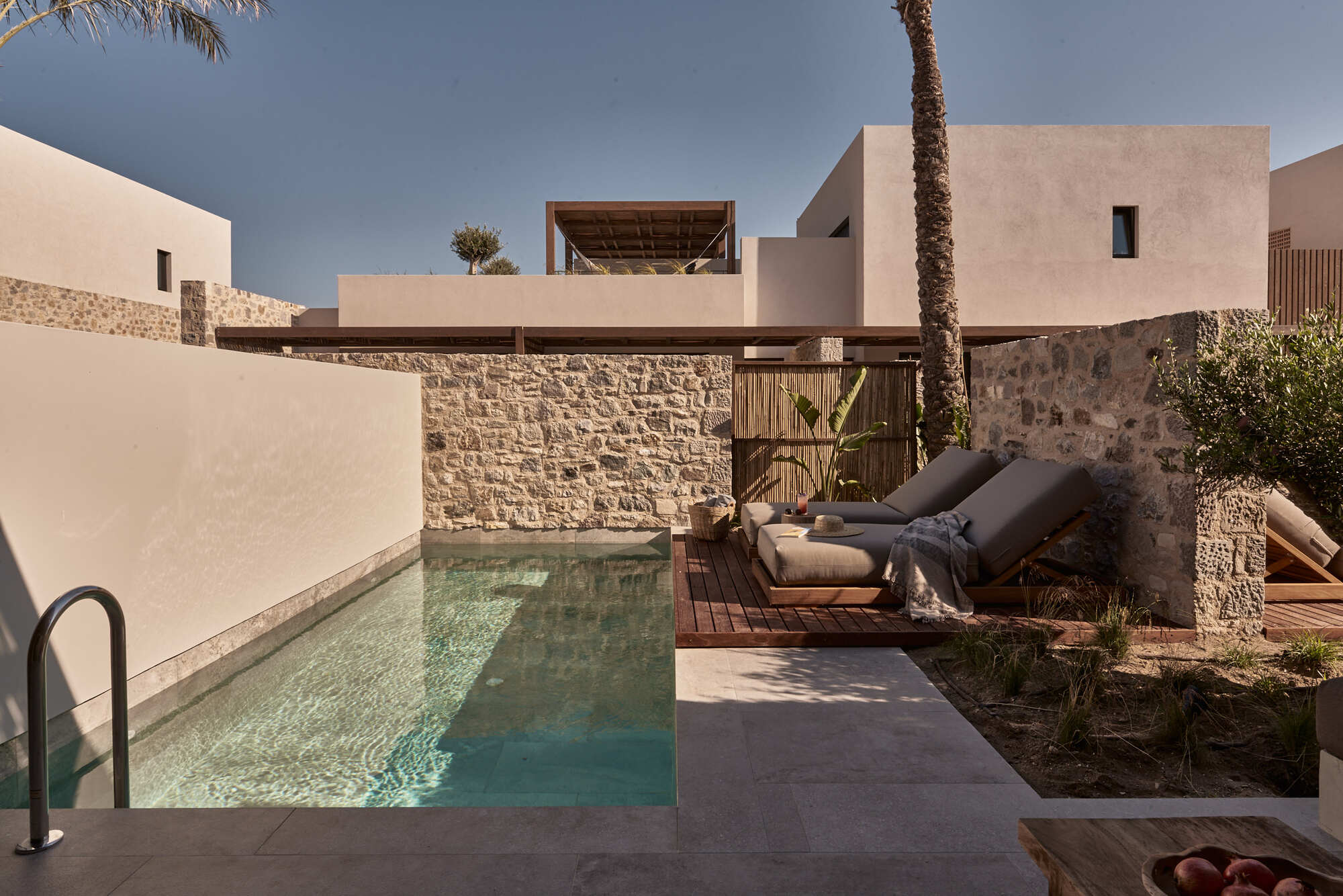 Private courtyard pool enclosed by stone walls with timber deck and loungers beside palm trees