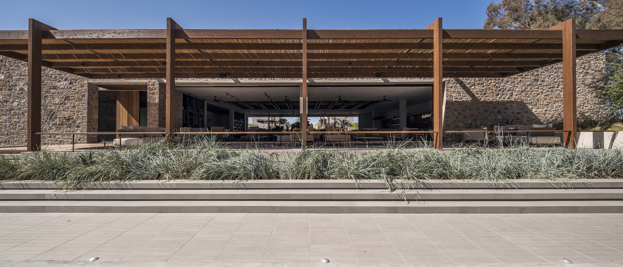 Stone facade with timber pergola and deep overhang fronting a planted bed of grasses