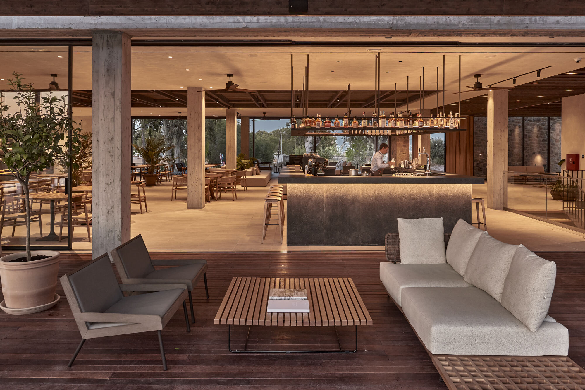 Open-air lounge and bar area with exposed concrete columns and timber ceiling under evening light