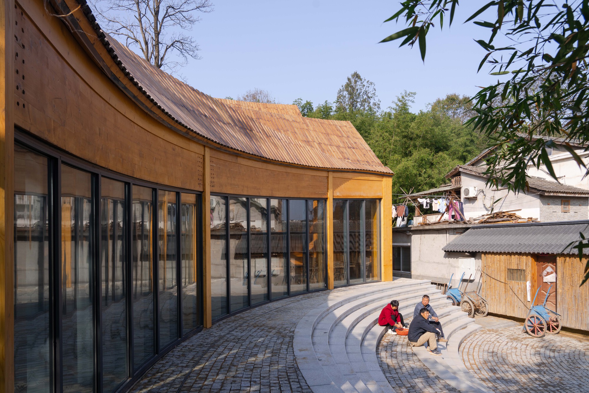 Curved corrugated metal roof over floor-to-ceiling glazing and paved courtyard with two seated figures