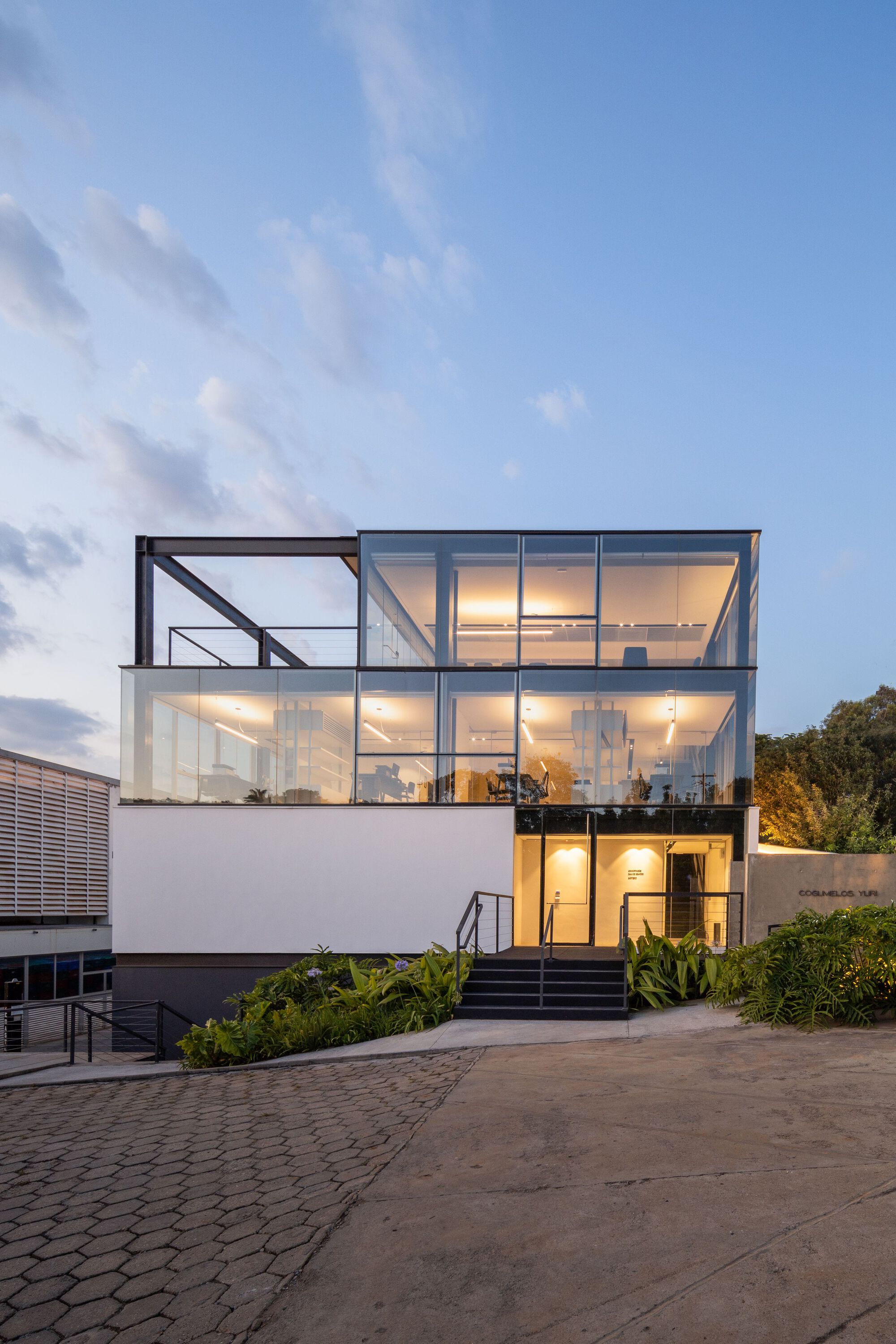 Front elevation of illuminated glass volume above white stucco base at twilight
