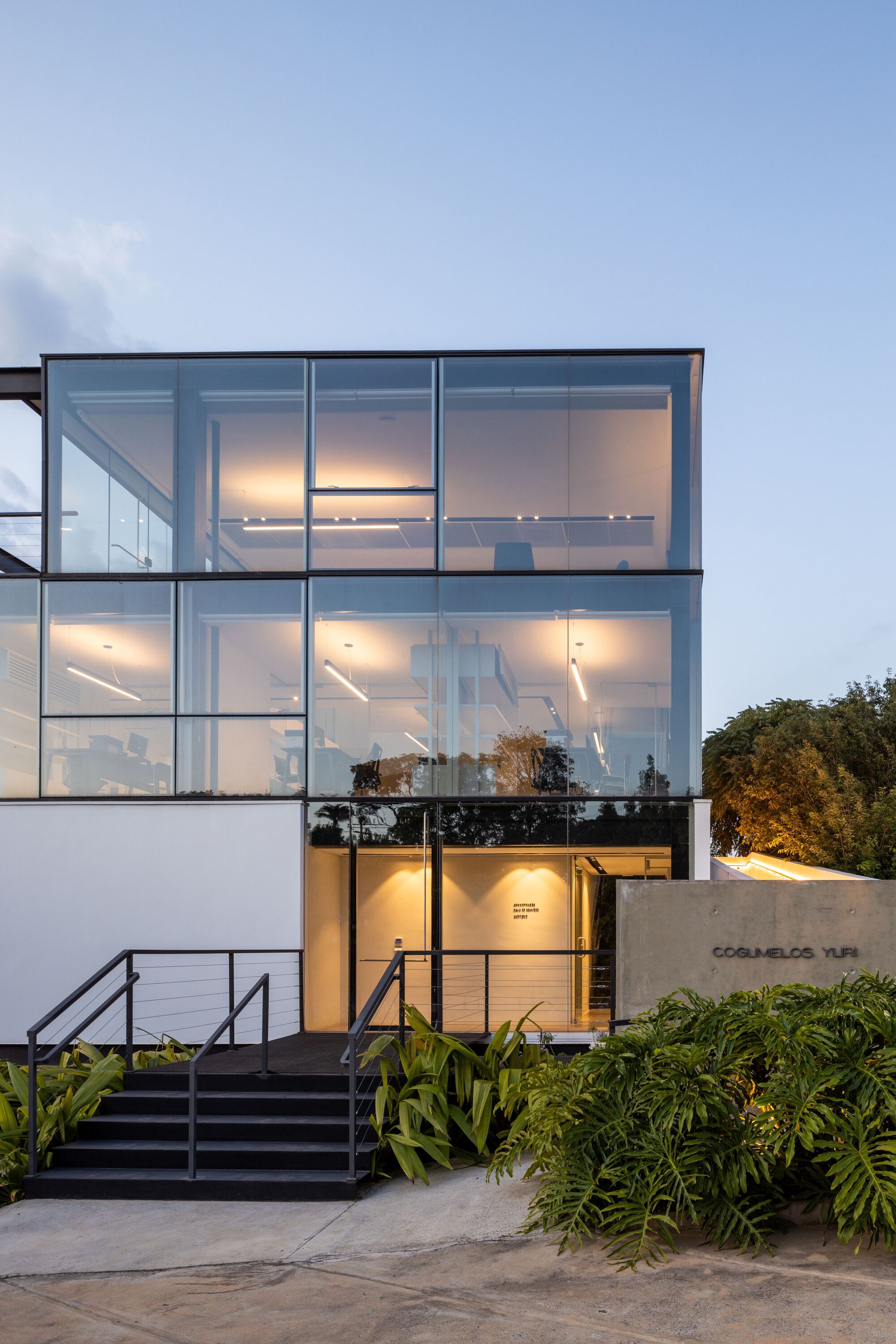 Three-story glass facade with exposed steel frame and interior lighting at dusk