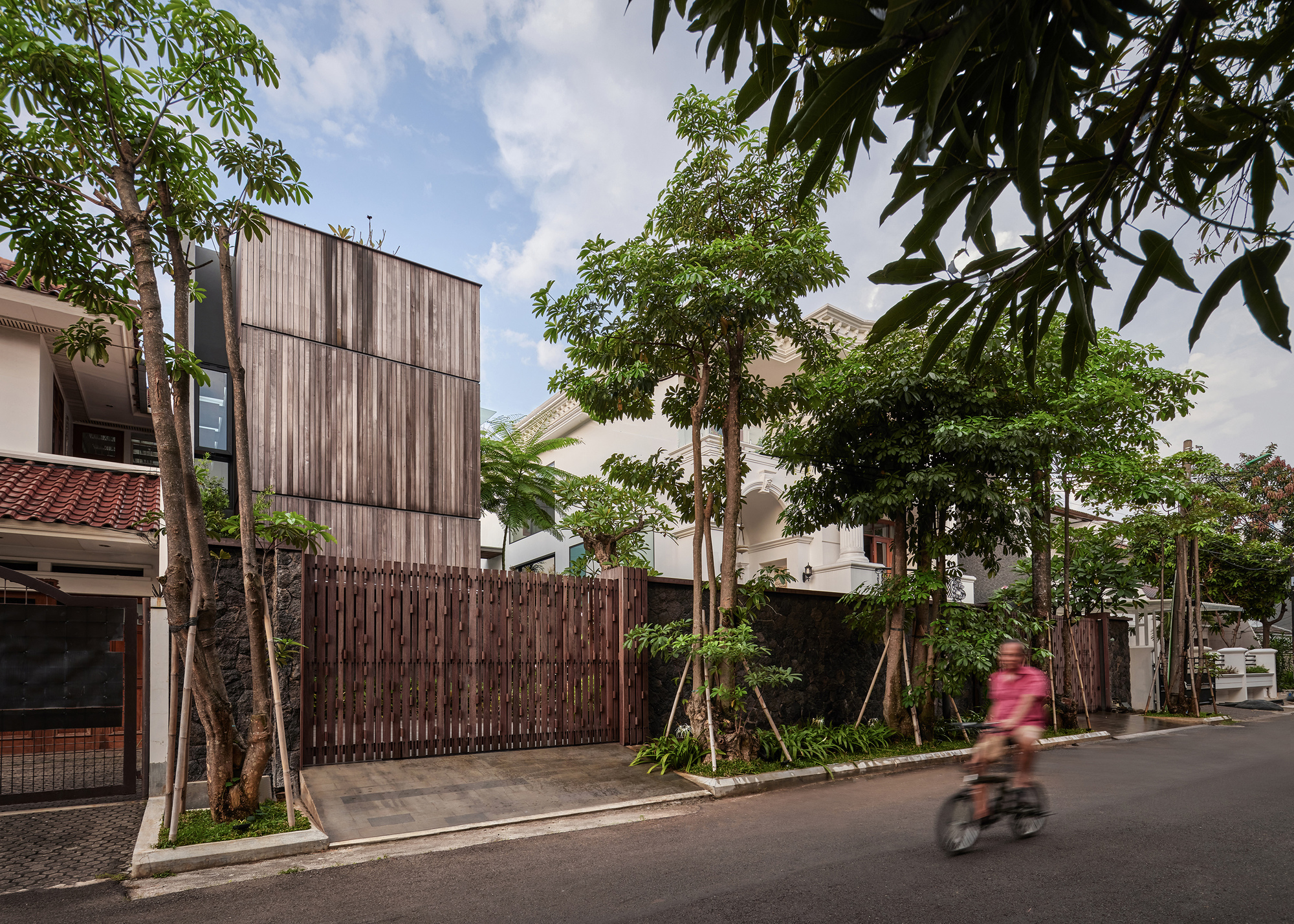 Street view of the vertical timber-clad volume behind a slatted wooden fence with a cyclist passing by