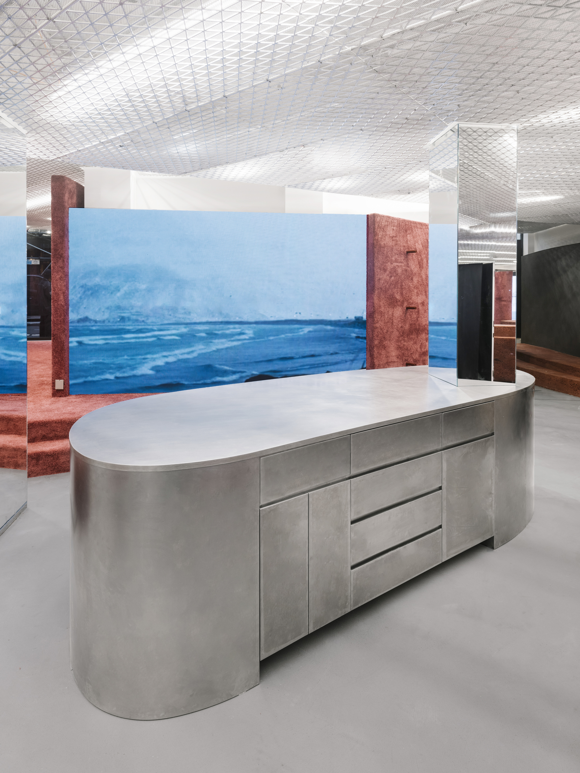 Oval stainless steel counter with integrated drawers against a large-scale ocean photograph wall mural