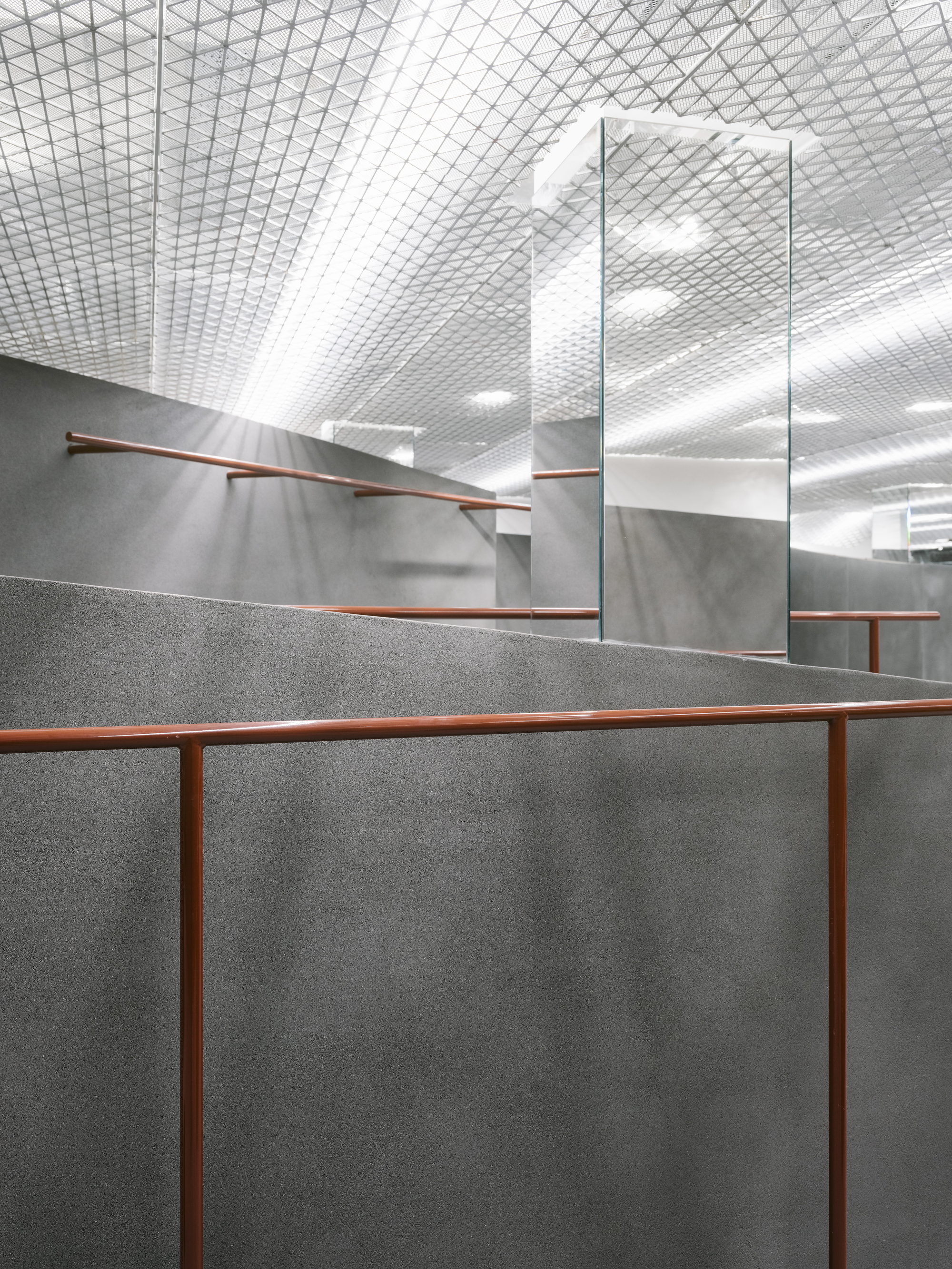 Interior view of grey concrete partition walls with copper handrails beneath a diamond-patterned mesh ceiling