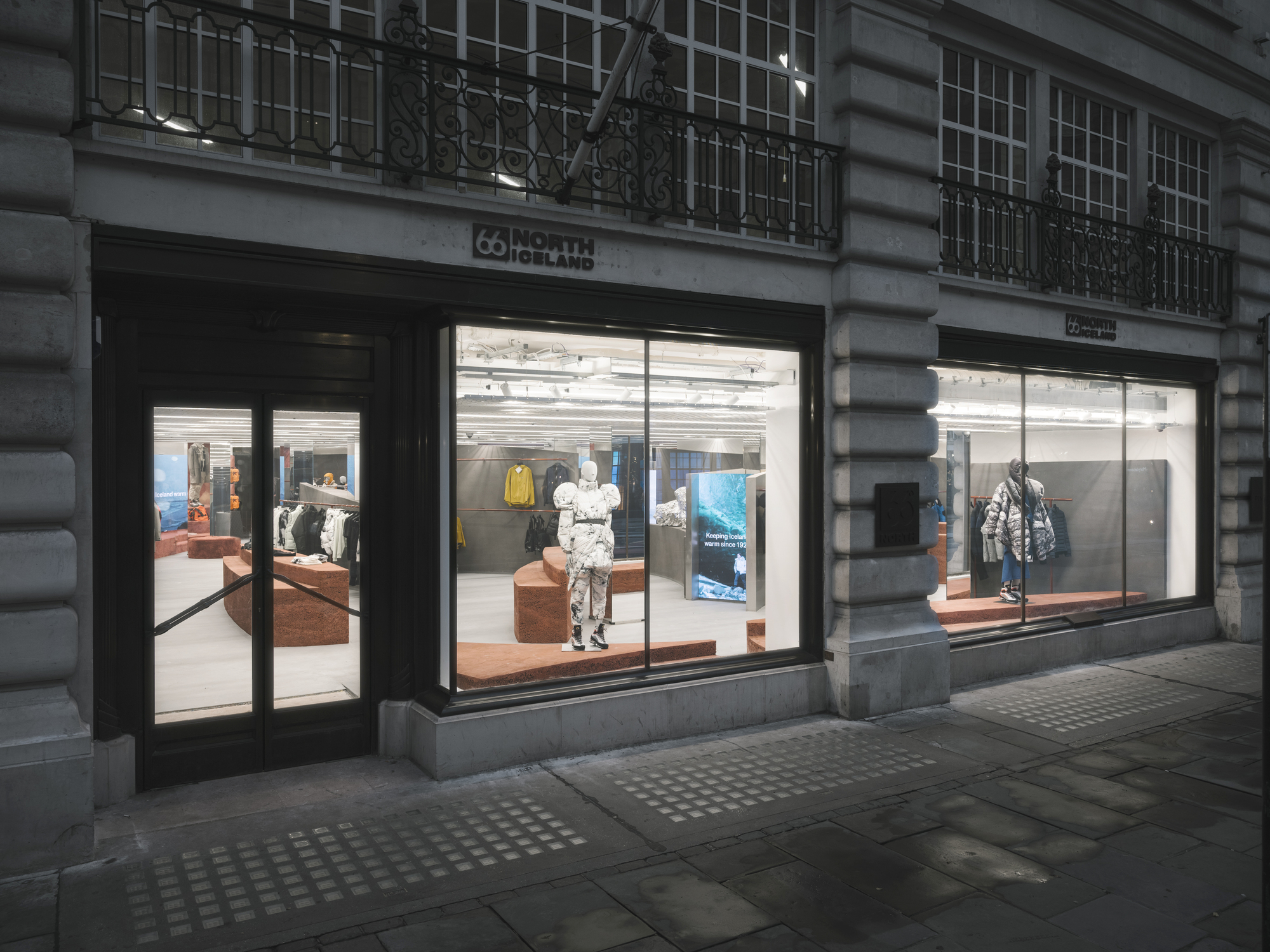 Evening view of symmetrical storefronts with illuminated interiors and mannequins beneath wrought iron balconies