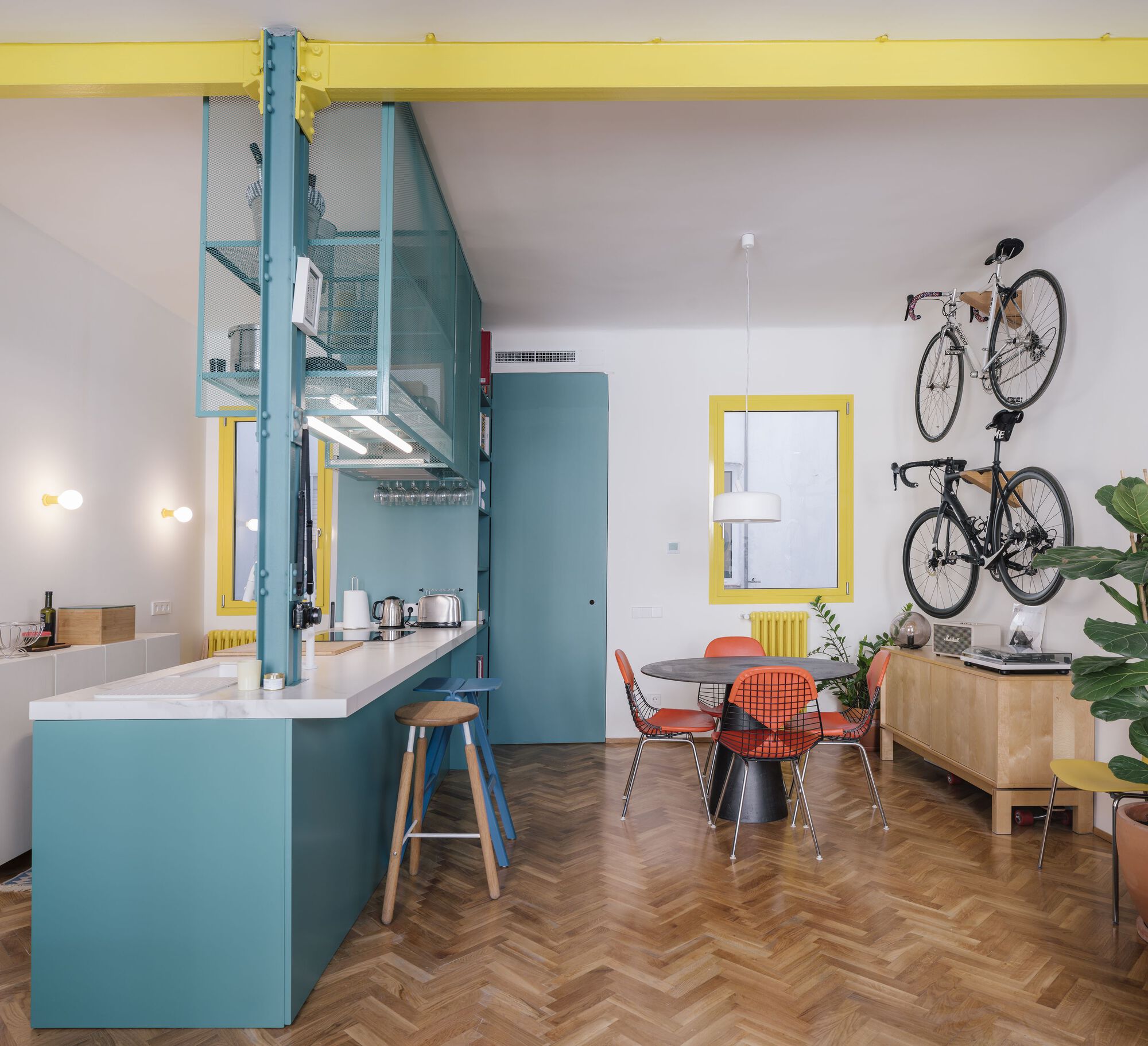 Open living area with teal kitchen island, yellow structural beam, and wall-mounted bicycles above herringbone flooring