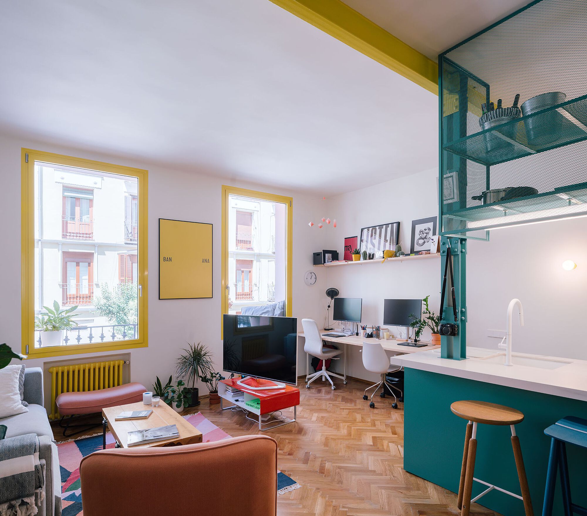 Combined living and work space with yellow-framed windows, teal storage wall, and herringbone wood floor