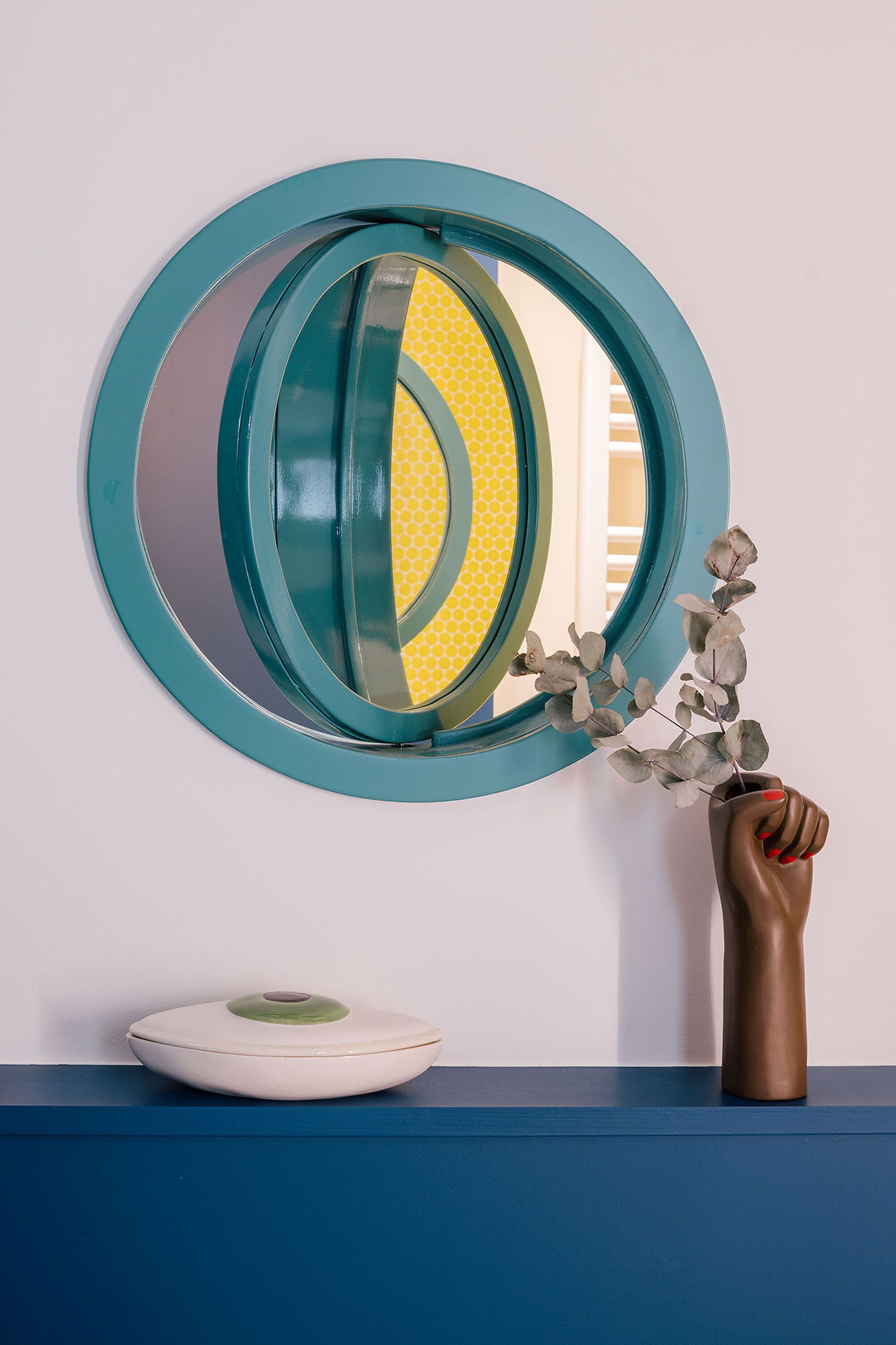 Circular porthole window with teal frame reflecting yellow interior detail and eucalyptus stem in vase below