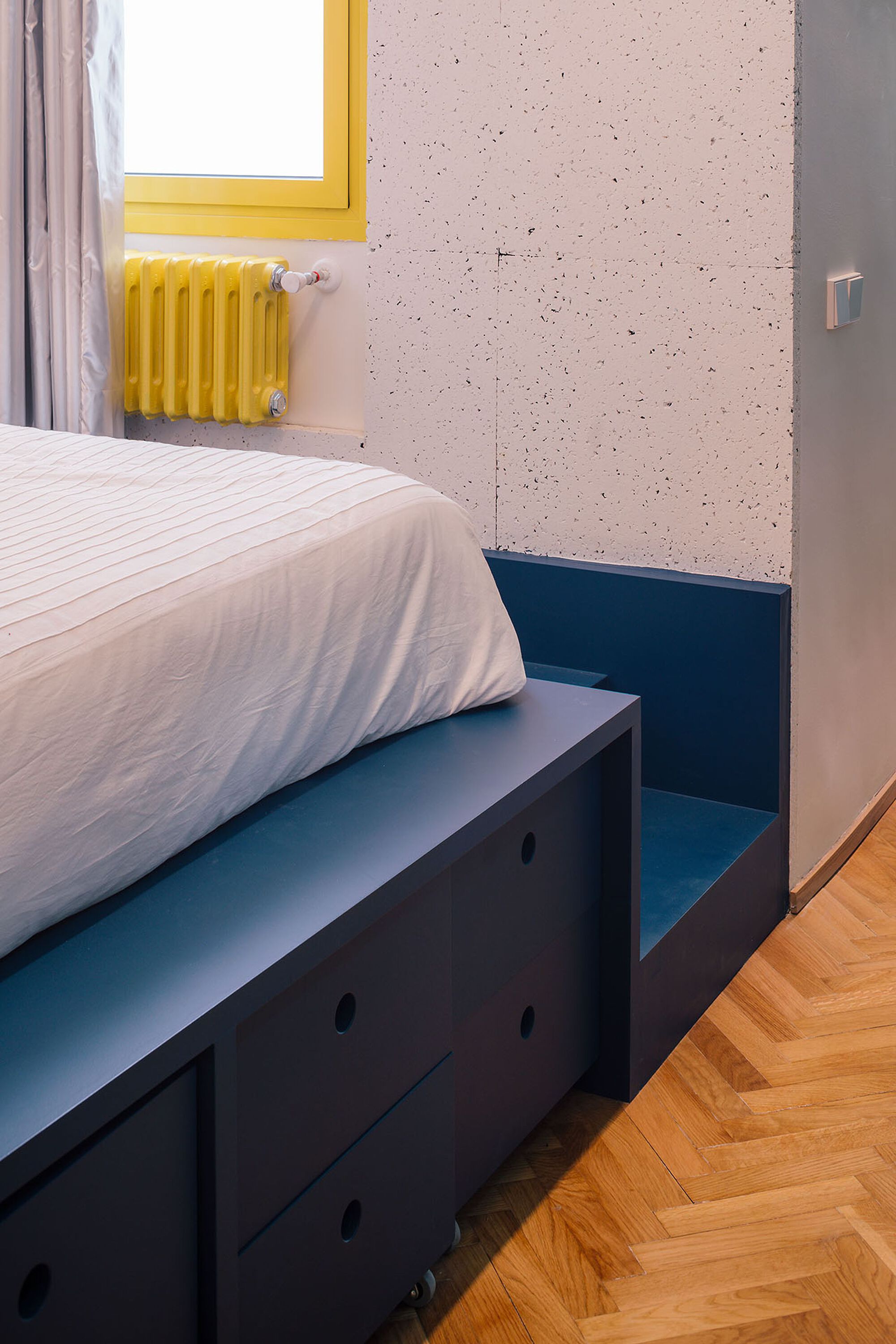 Bedroom corner with blue platform bed against textured pink wall and yellow radiator beneath window