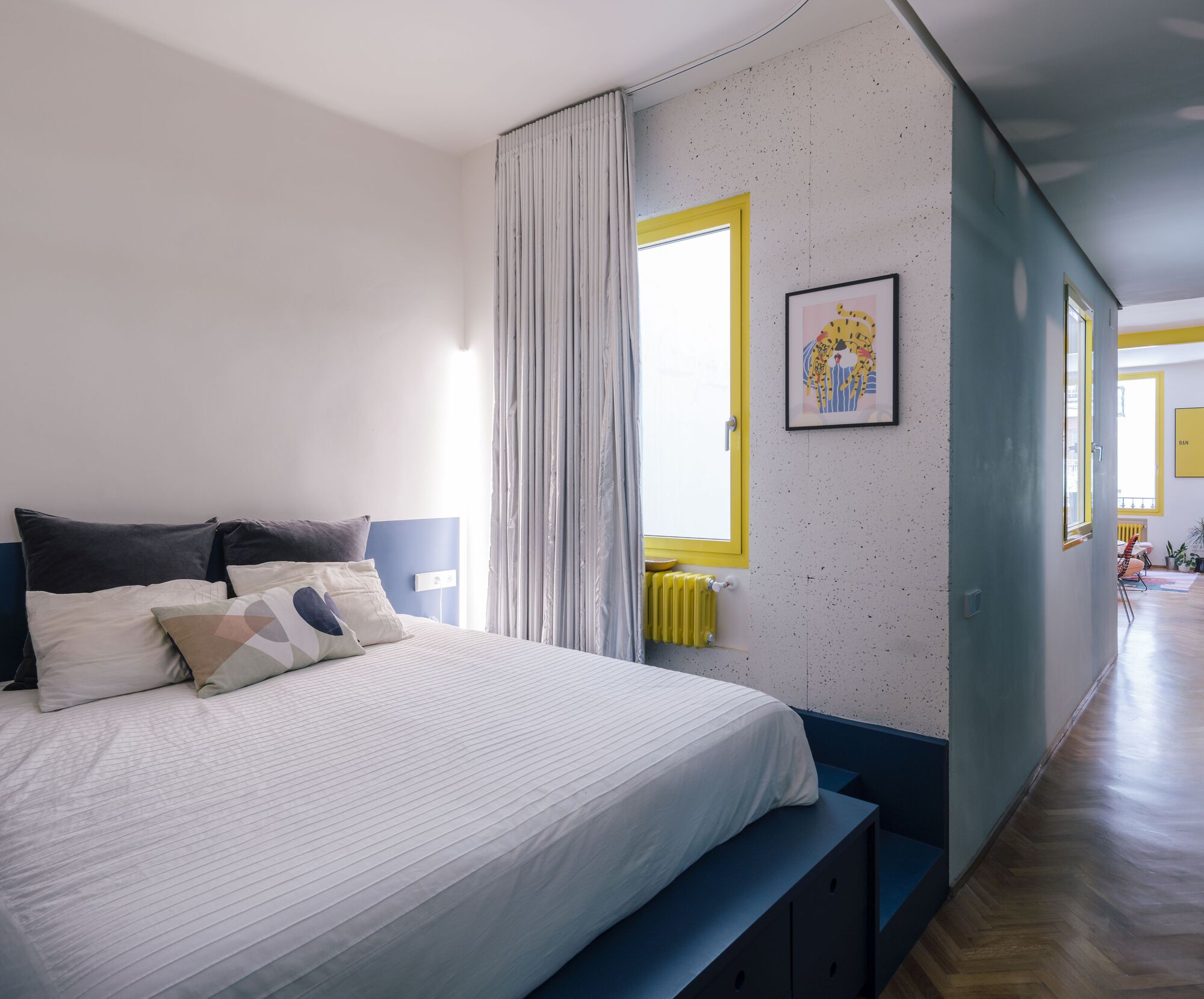 Bedroom with exposed concrete wall, yellow-framed window and yellow radiator beneath sheer curtains