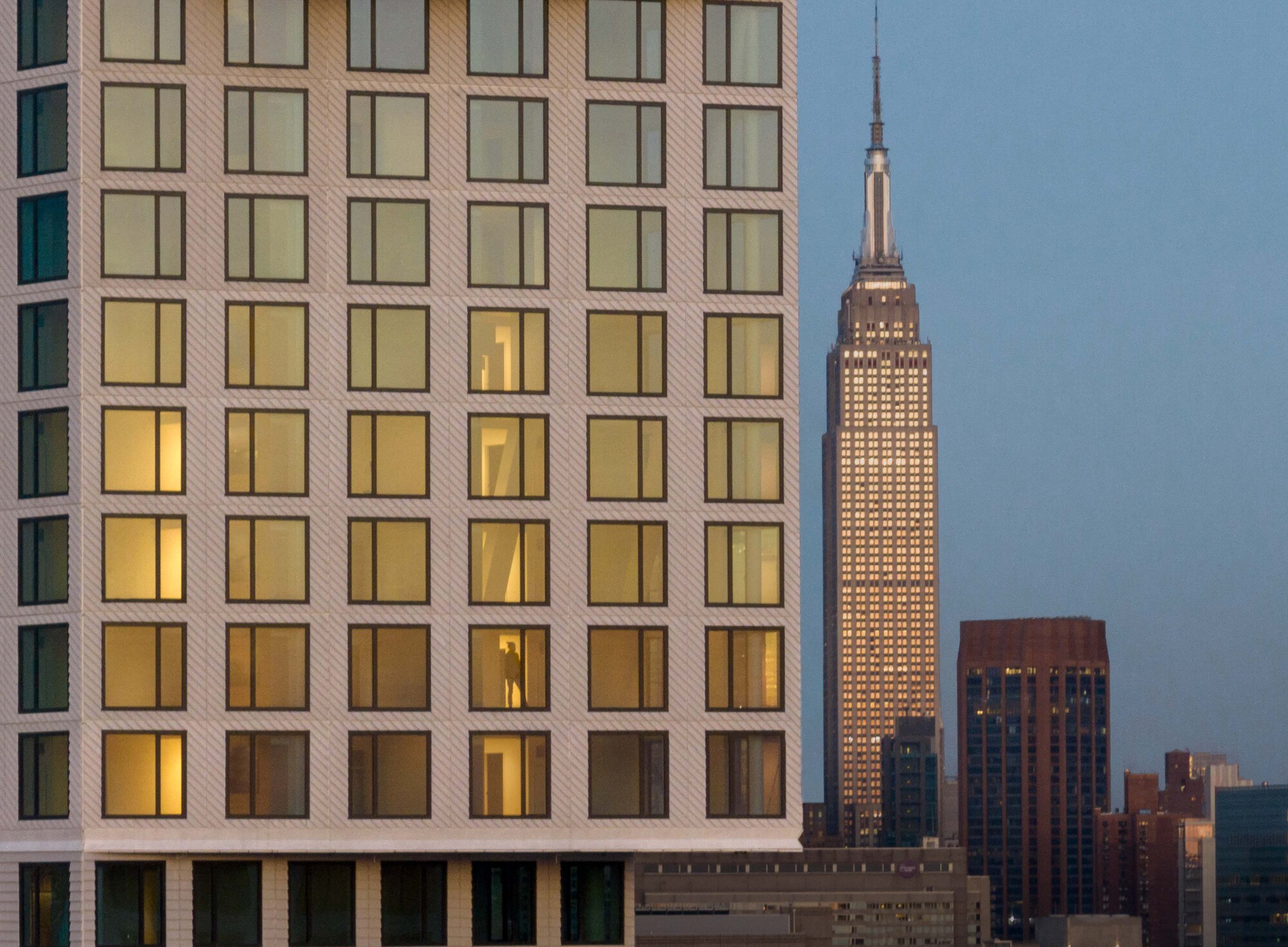 Residential tower facade with illuminated windows at dusk and a historic skyscraper in the background