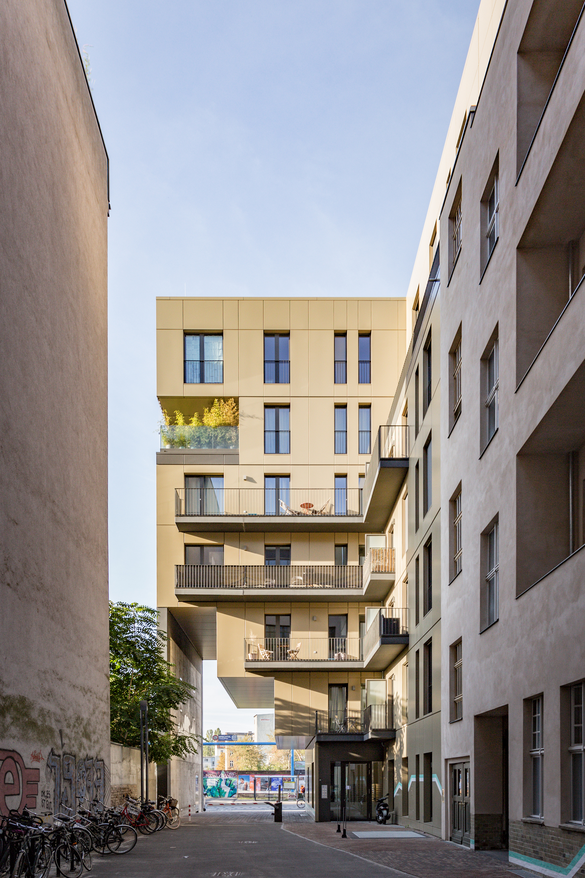Street view framed by adjacent buildings showing the golden-paneled facade with protruding balconies and bicycles below