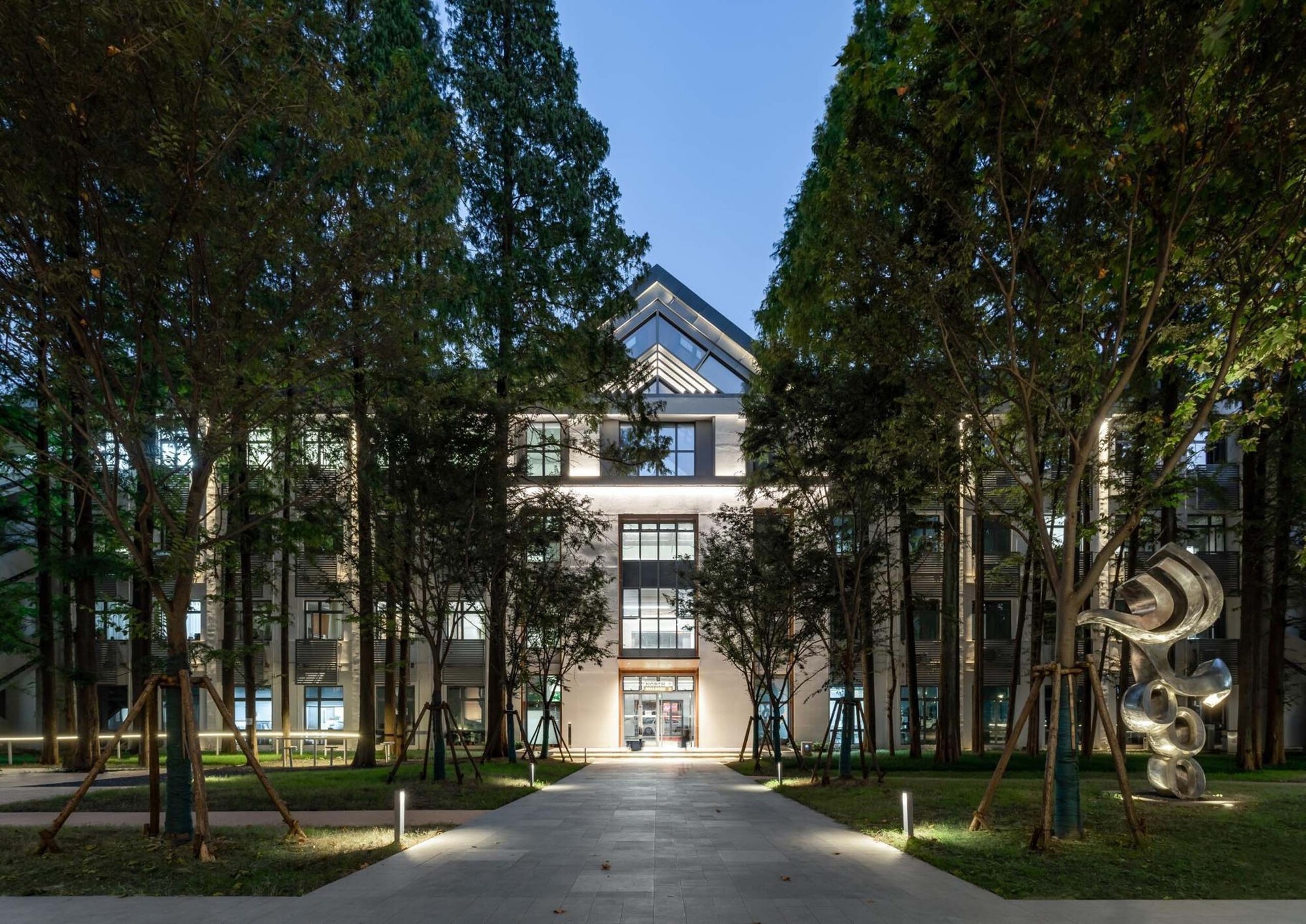 Front facade illuminated at dusk, framed by rows of tall trees along a paved walkway