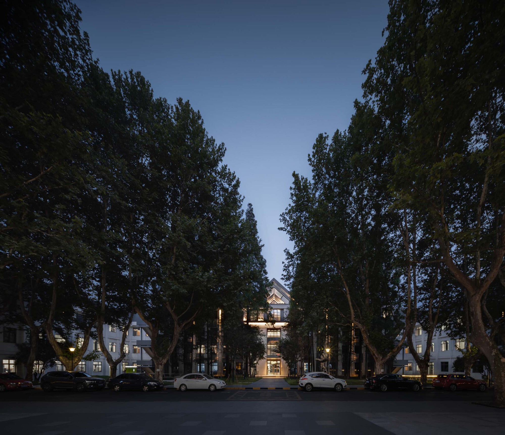 Illuminated central building framed by mature trees and parked cars in evening light