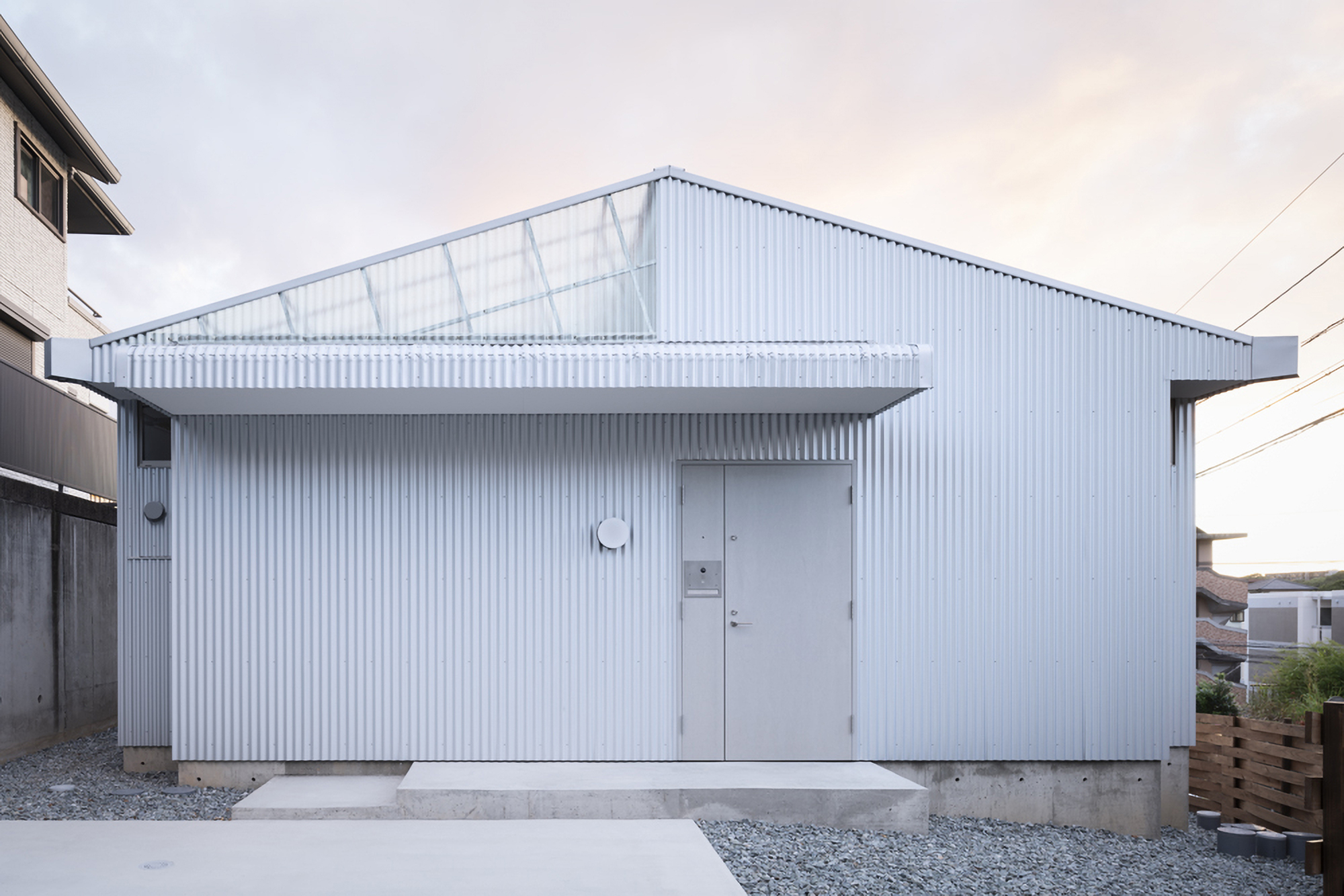 Street view of the white corrugated metal facade with a central door and translucent roof panels