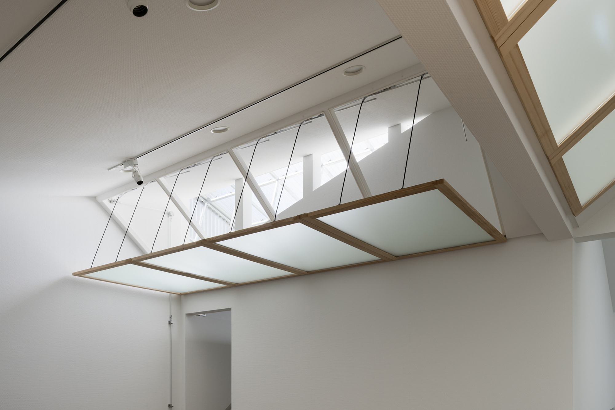Diagonal lattice glazing system above a suspended timber-framed panel in a white interior