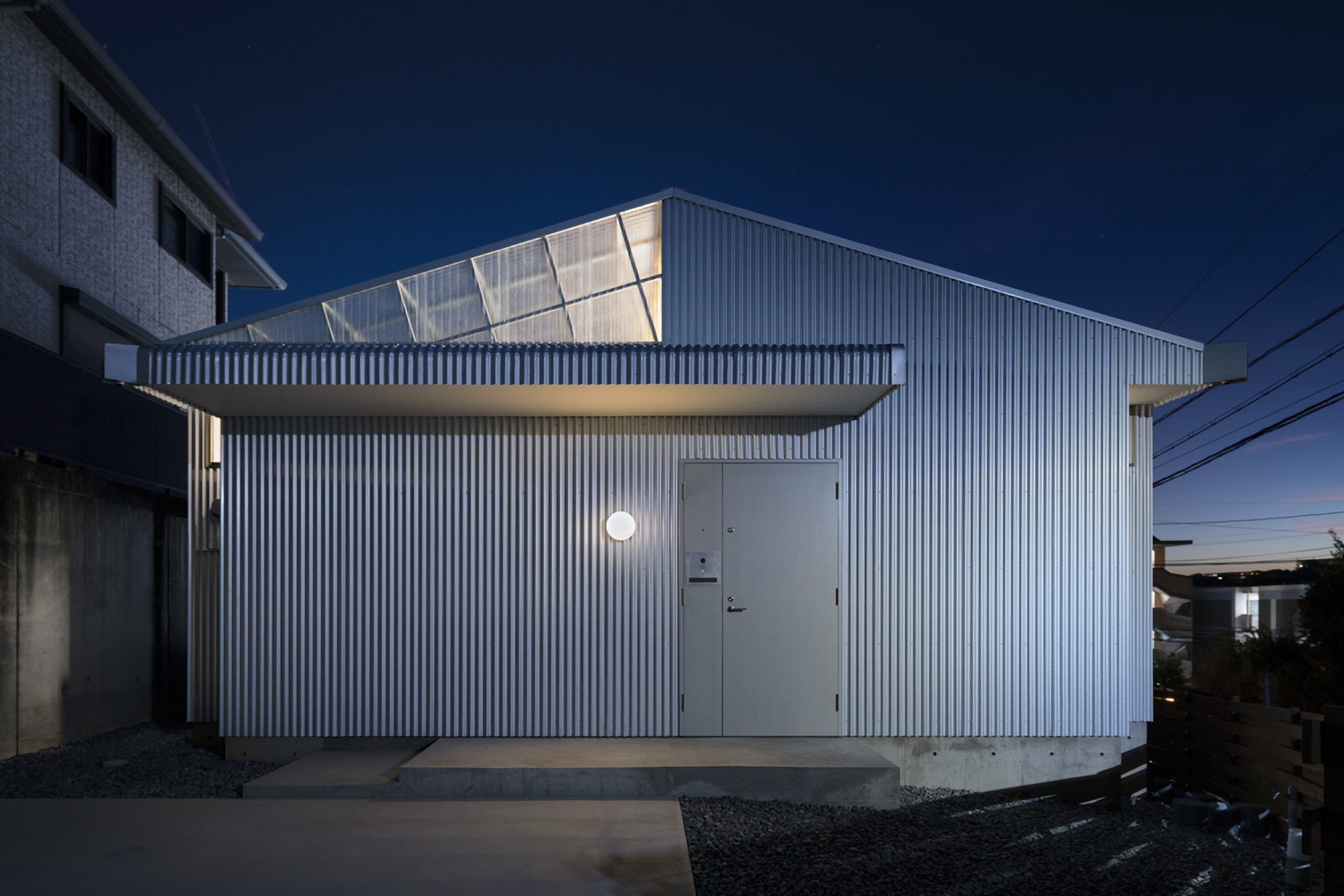 Corrugated metal facade with illuminated sawtooth roof and entry door at twilight