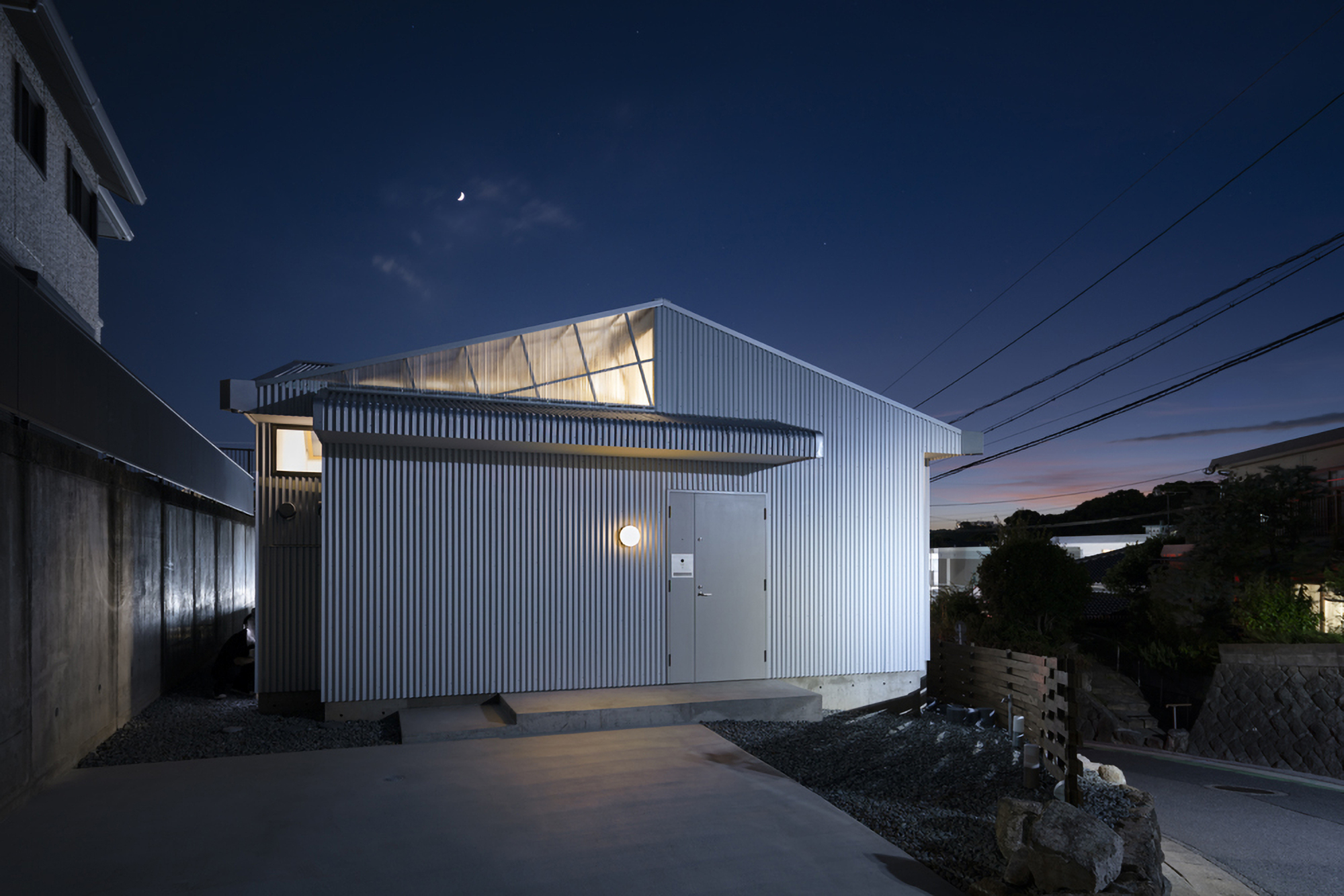 Street view of the vertical corrugated cladding and glowing clerestory windows under a crescent moon