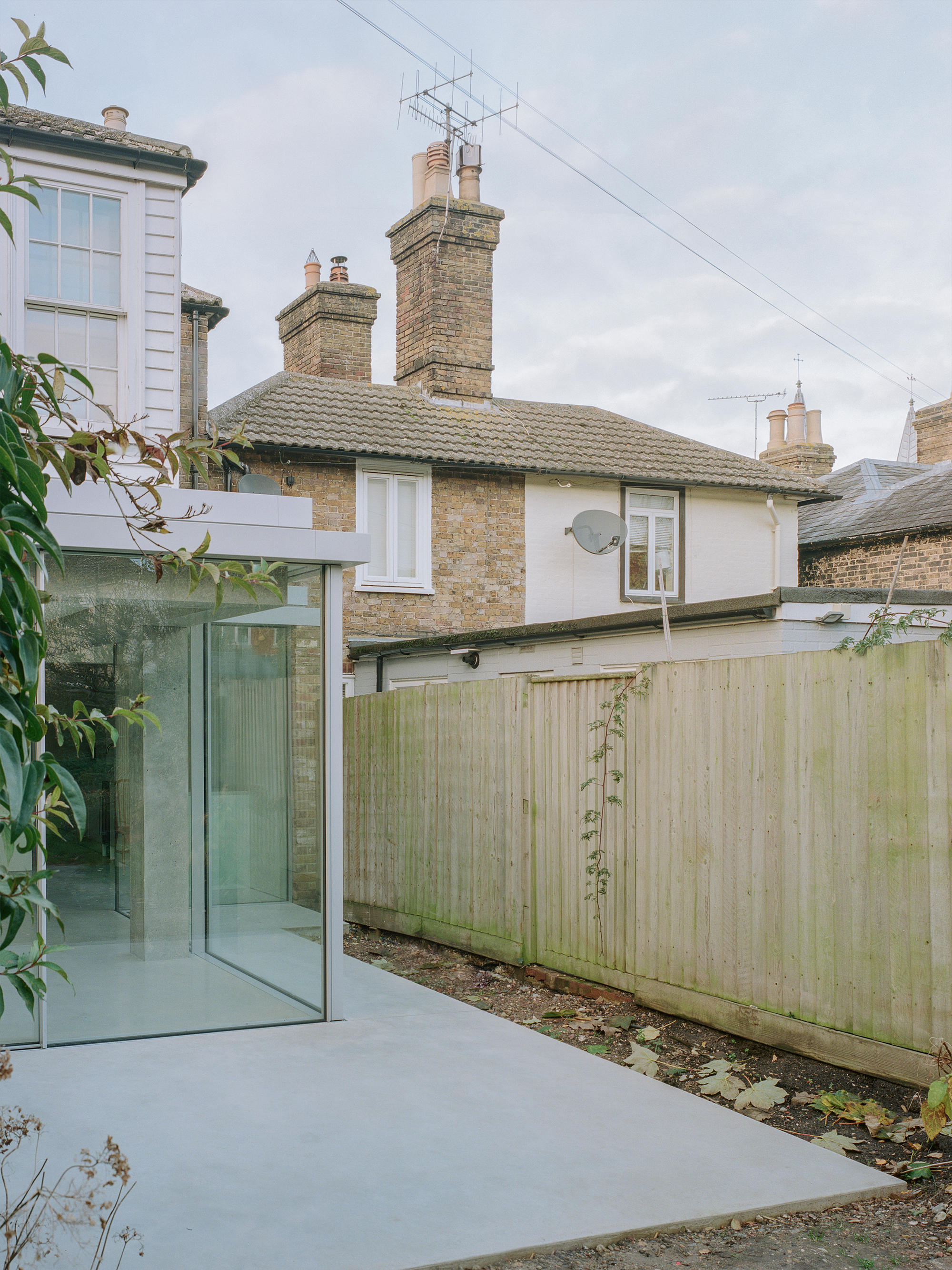 Rear extension with flat concrete roof and glazed corner beside weathered timber fence and existing brick houses